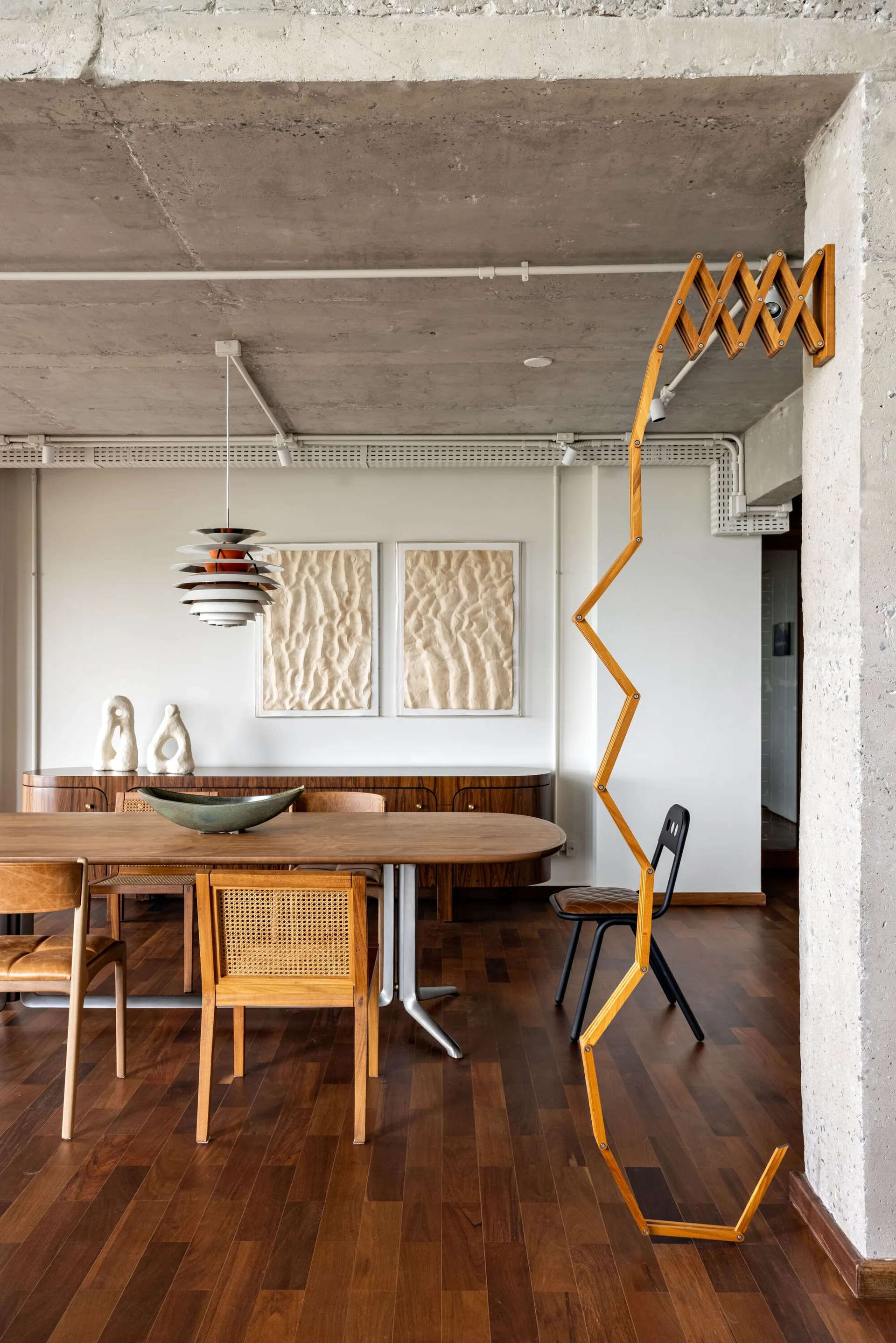 A modern dining room features an oval wooden dining table, cane-back chairs, a curved sideboard, and a raw concrete ceiling.