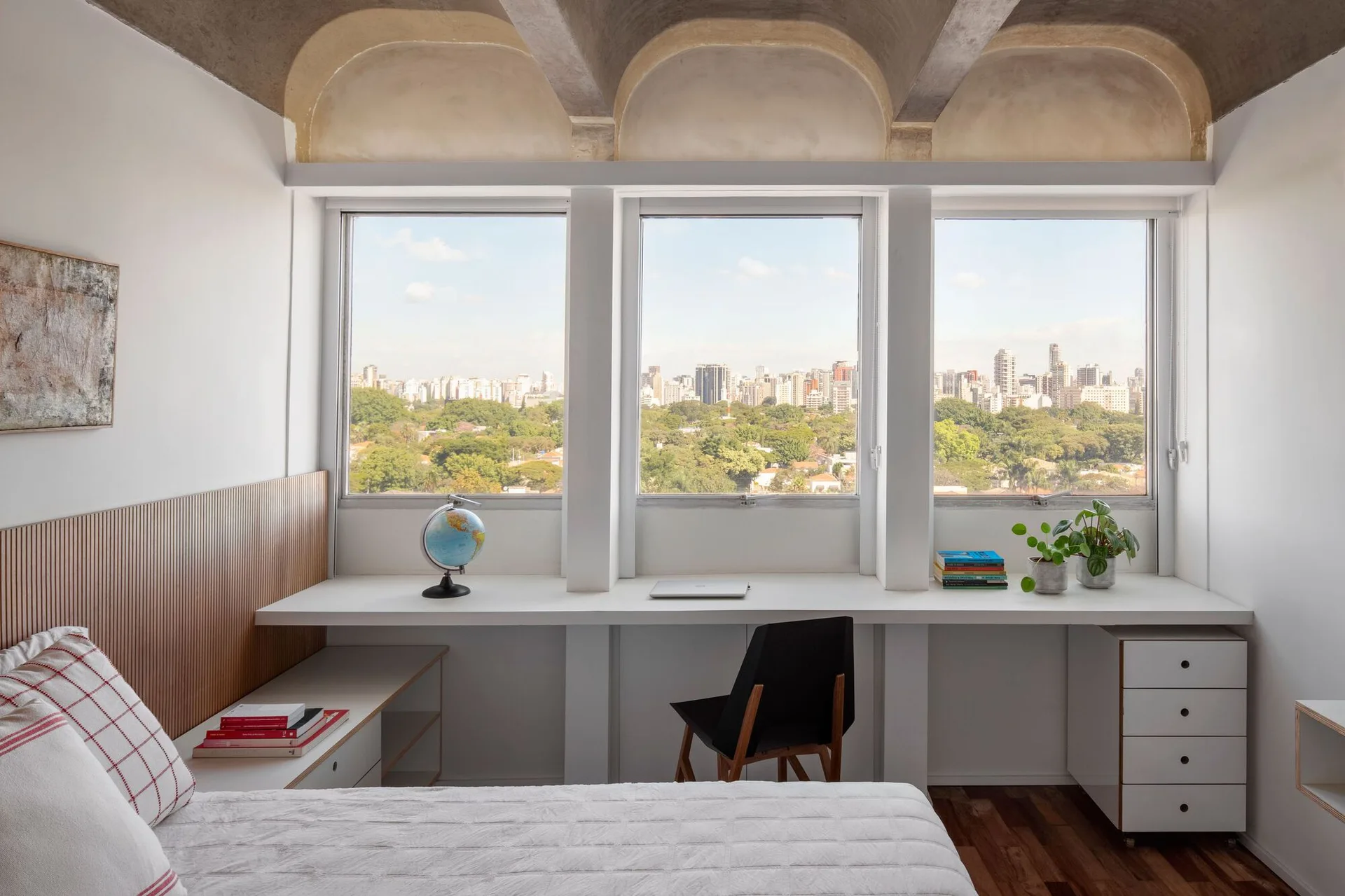 A modern bedroom features a white built-in desk under tripartite windows overlooking a city skyline, with a vaulted concrete ceiling above.