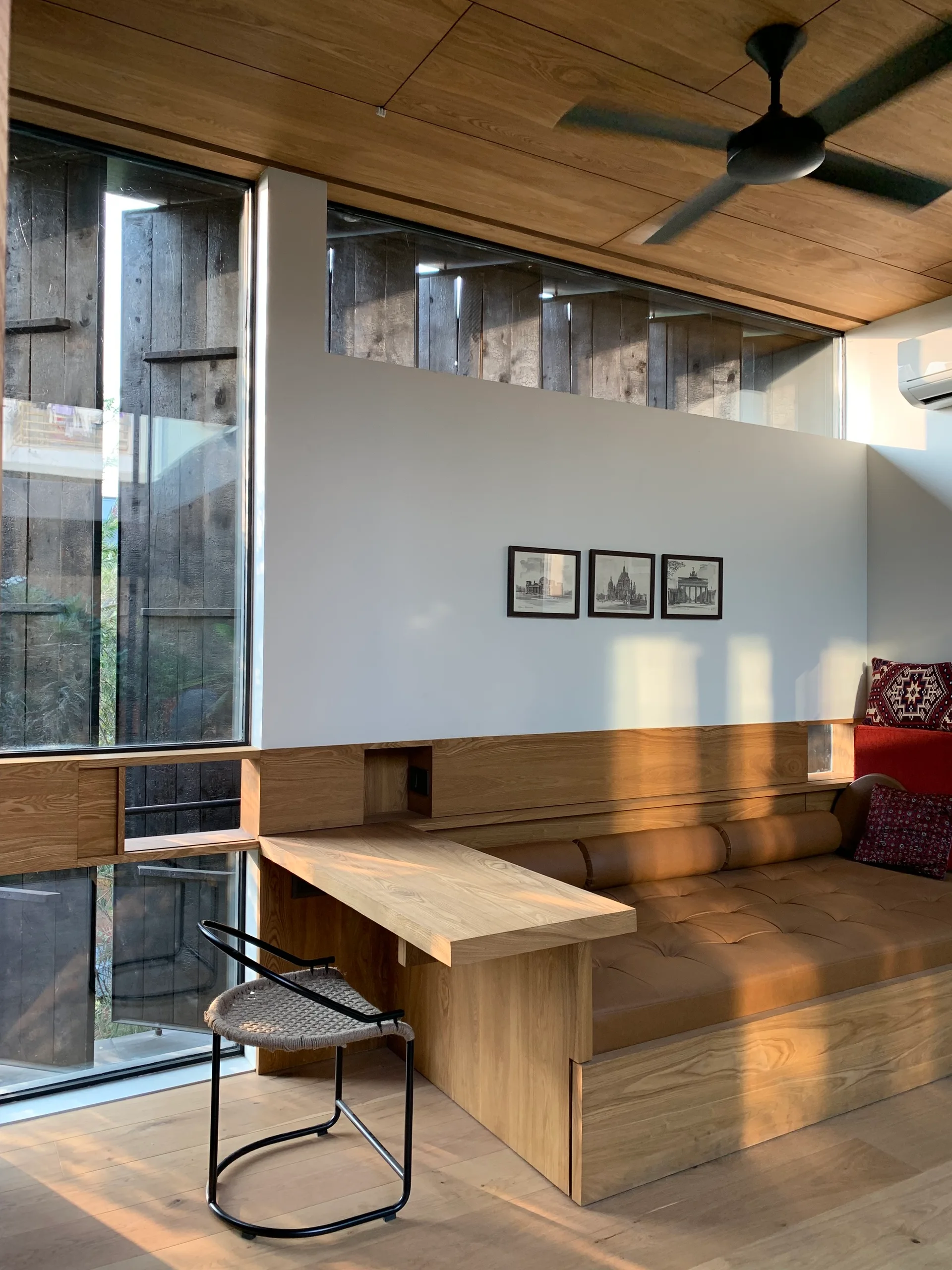A modern room featuring a built-in wooden daybed with a tan leather cushion, an integrated wooden desk, and large glass windows.