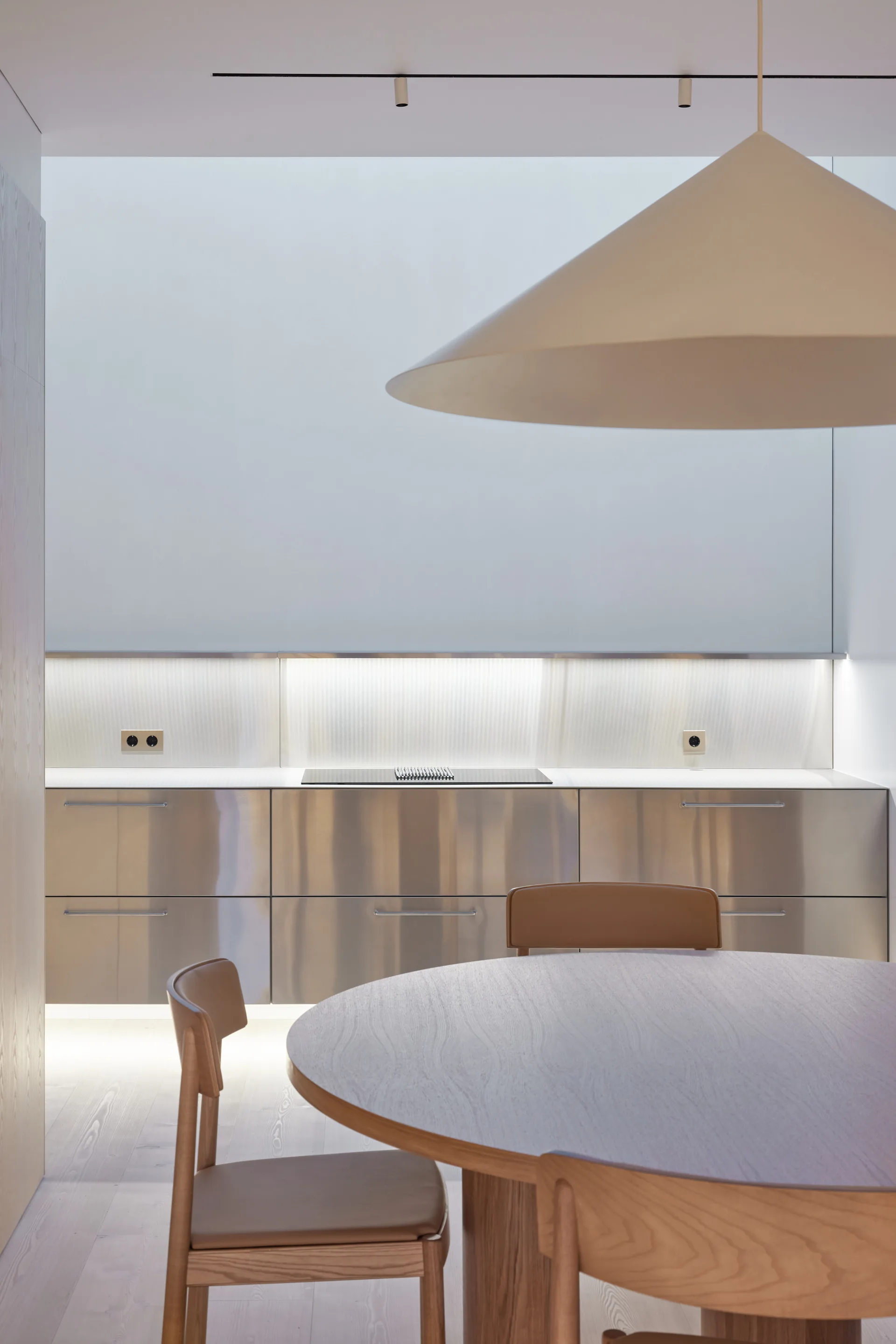 A modern kitchen featuring brushed stainless steel cabinets, a ribbed glass backsplash with integrated lighting, and a round wooden dining table.