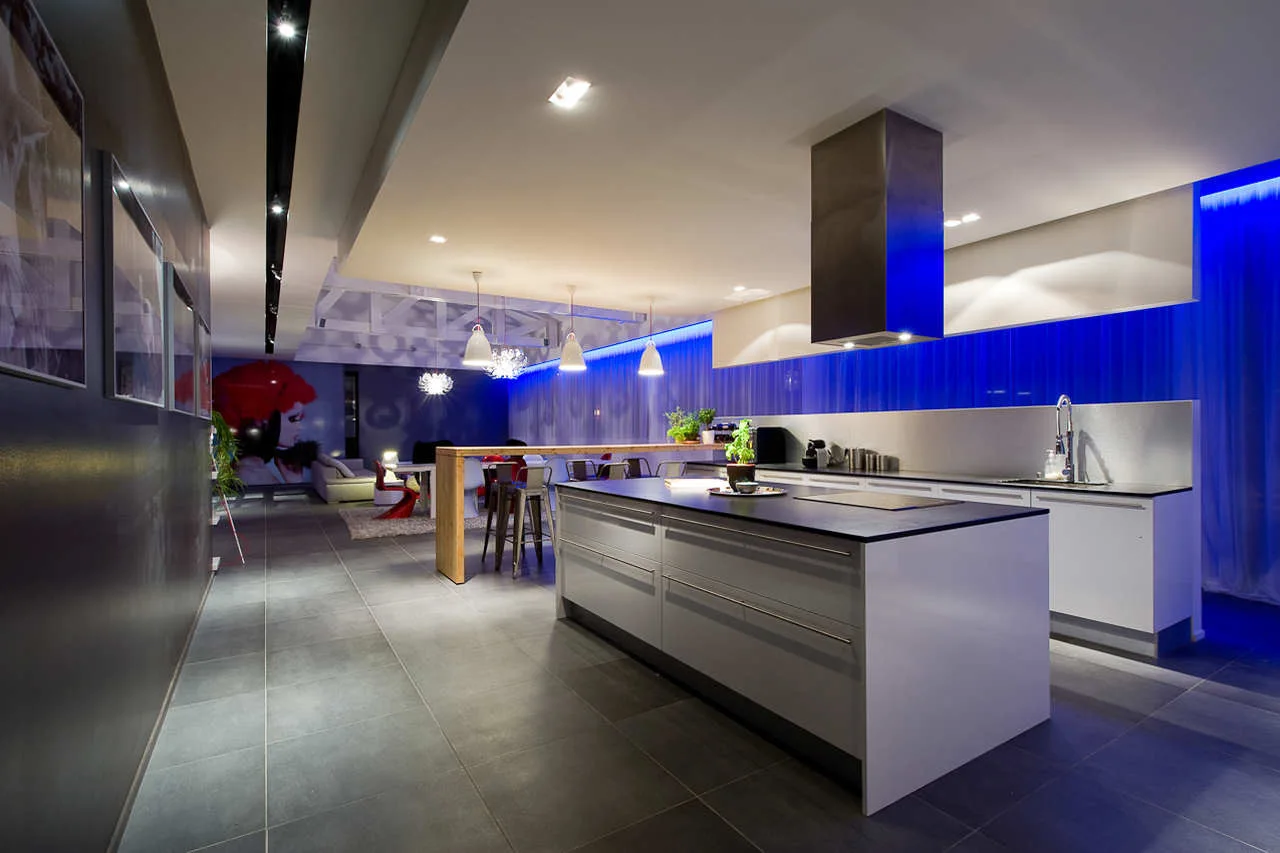 A modern open-concept kitchen featuring a large grey island, white cabinetry, a stainless steel range hood, and bright blue LED wall lighting.