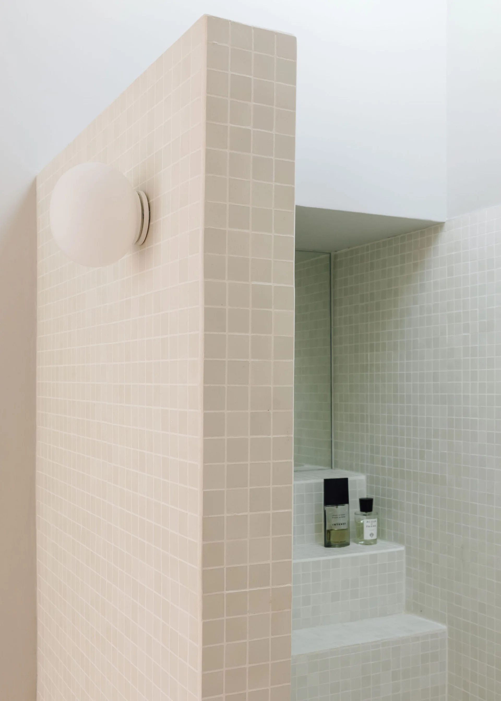 A minimalist bathroom features a partition wall covered in beige square mosaic tiles, a globe wall sconce, and a stepped tiled vanity ledge.
