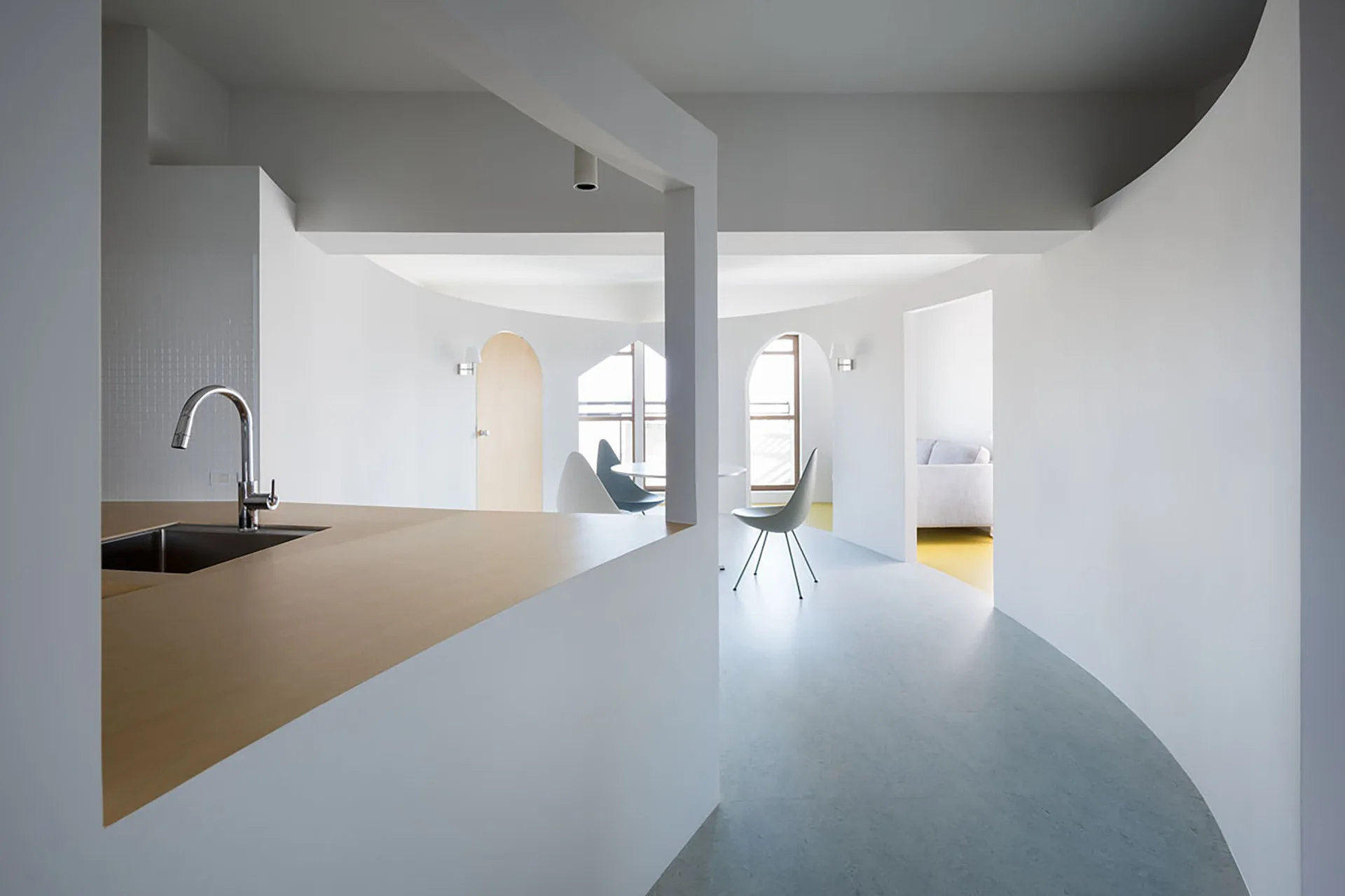 A minimalist open-plan kitchen featuring a light wood island with a sink, curved white walls, and a dining space in the background.