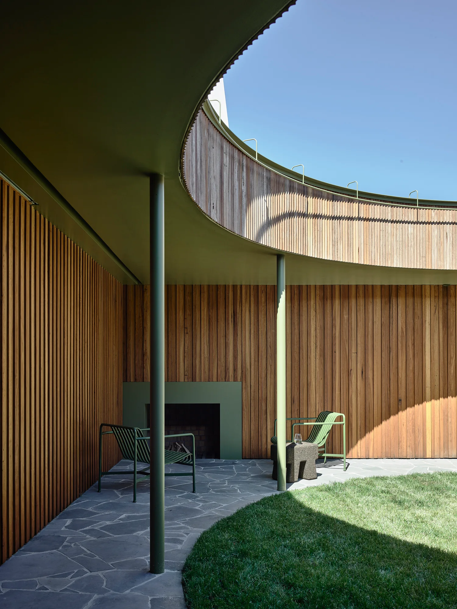 A curved outdoor patio features vertical wood-slat walls, two green metal lounge chairs near an inset fireplace, and irregular stone paving.
