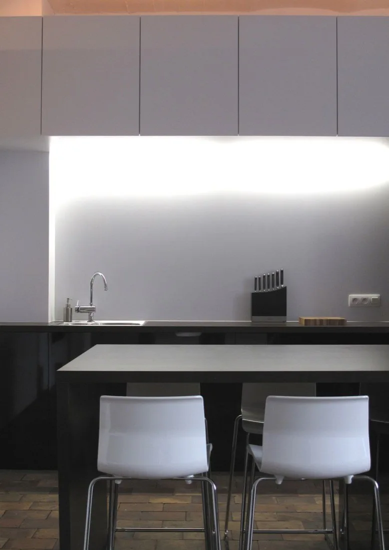 A minimalist kitchen features matte grey upper cabinets, bright under-cabinet lighting, a black counter, and white modern bar stools.