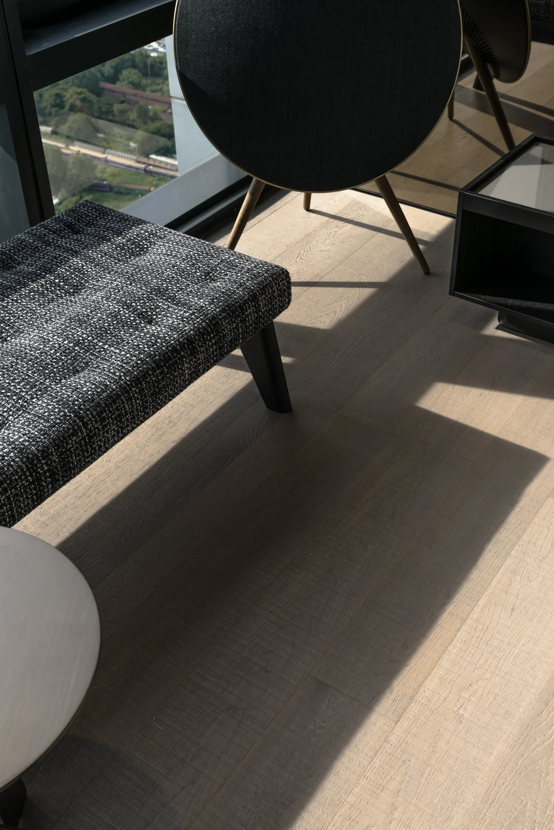 A textured black and white bench and a circular black chair on light wood floors next to a large window casting geometric shadows.