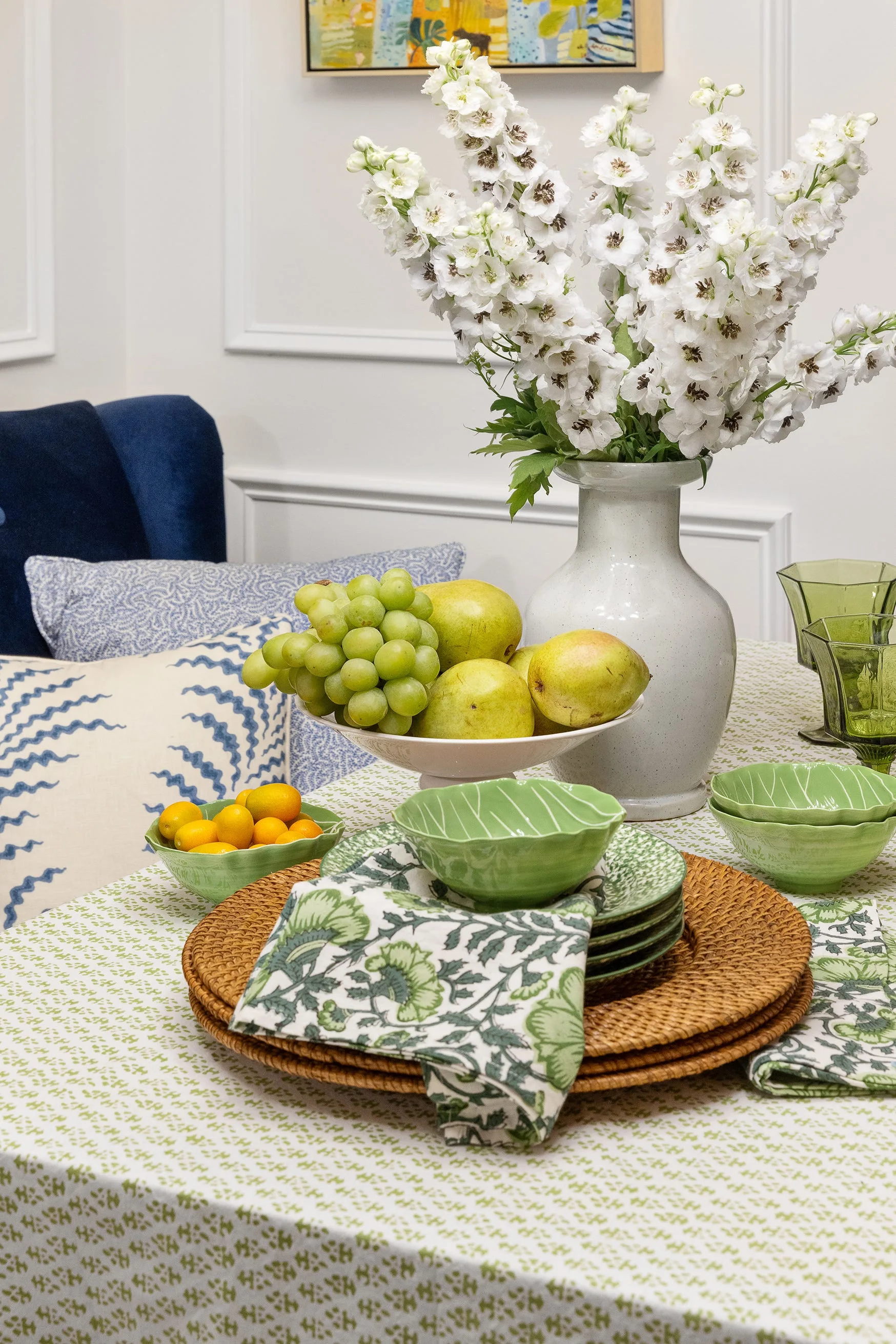 A close-up of a dining table styled with a green and white floral tablecloth, rattan charger plates, green ceramic bowls, and white flowers in a vase.
