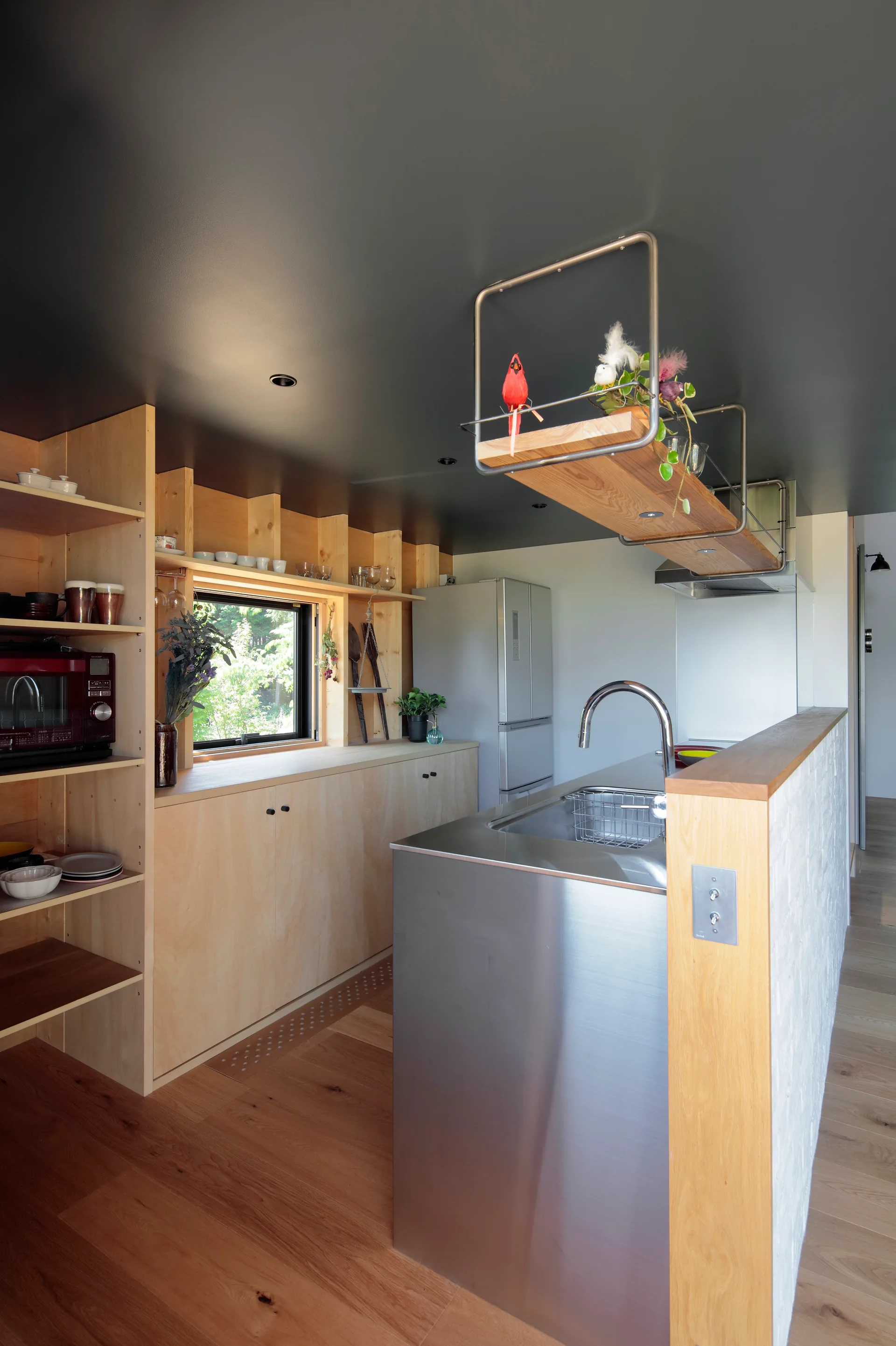 A minimalist kitchen featuring light wood cabinetry, a stainless steel island with a sink, a suspended shelf, and a dark grey painted ceiling.