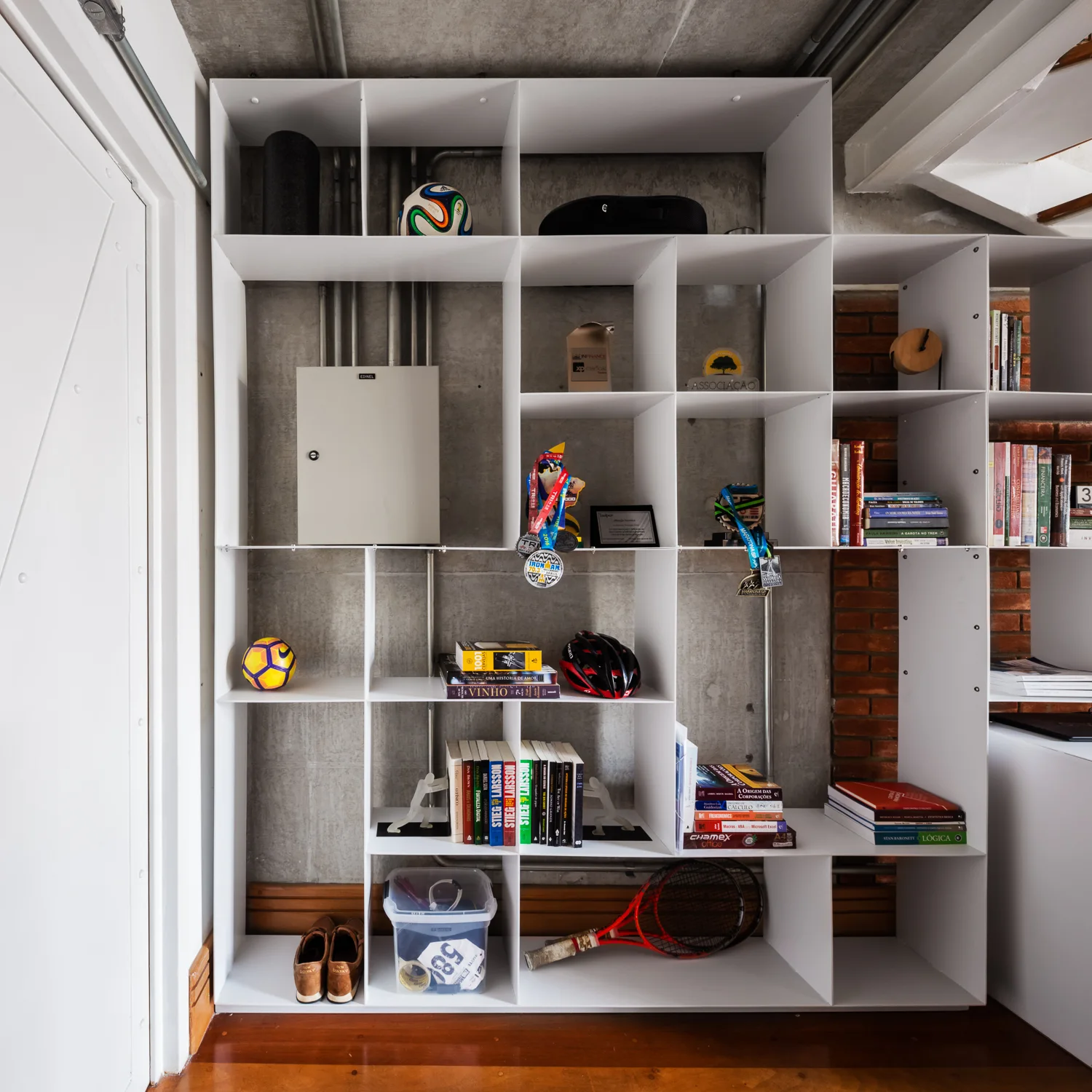 A white open-grid bookshelf filled with books and sports gear sits against an exposed concrete and red brick wall.