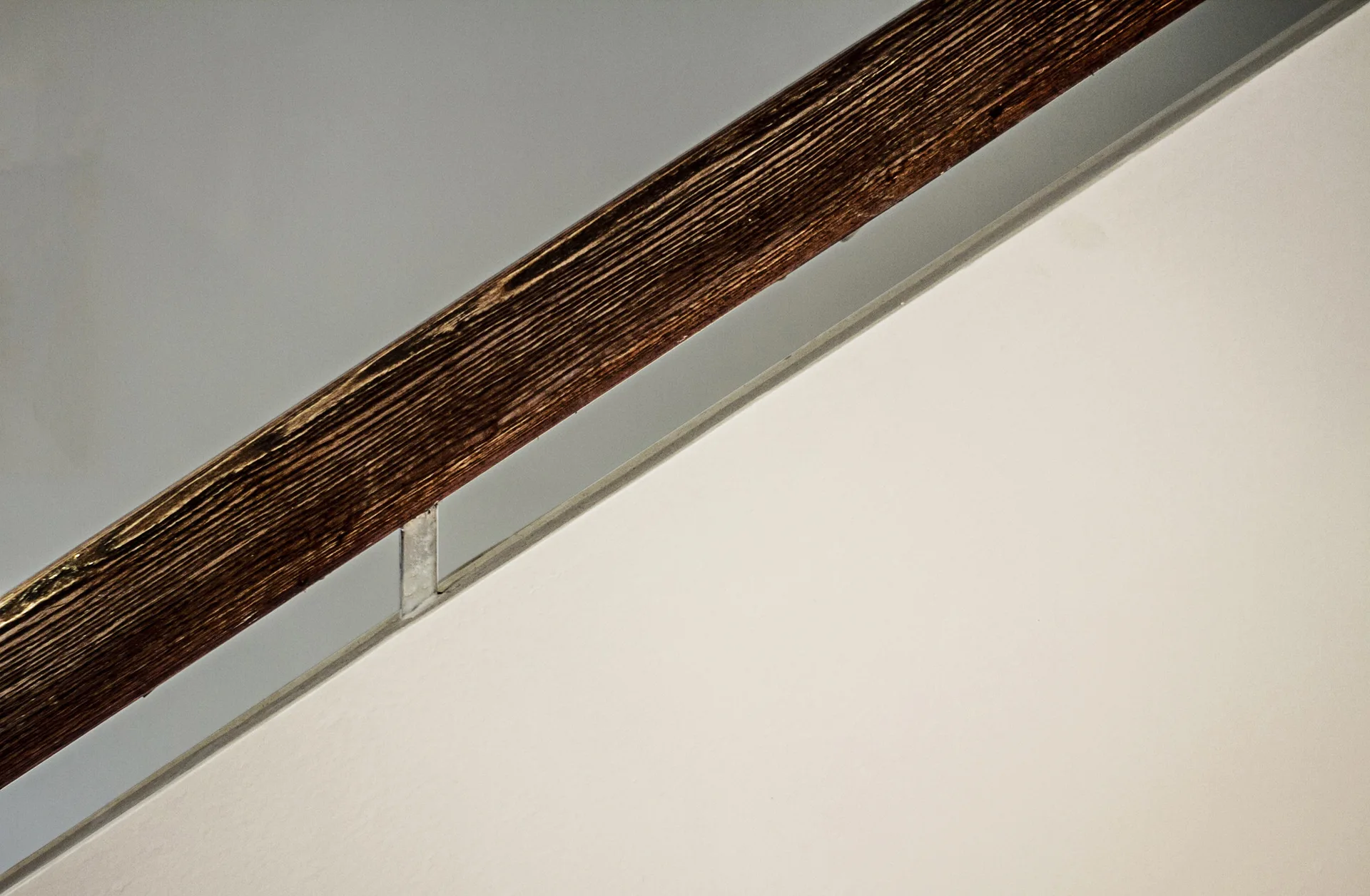 A close-up of a diagonal dark wood handrail with prominent grain texture mounted to a plain white wall with a thin metal bracket.