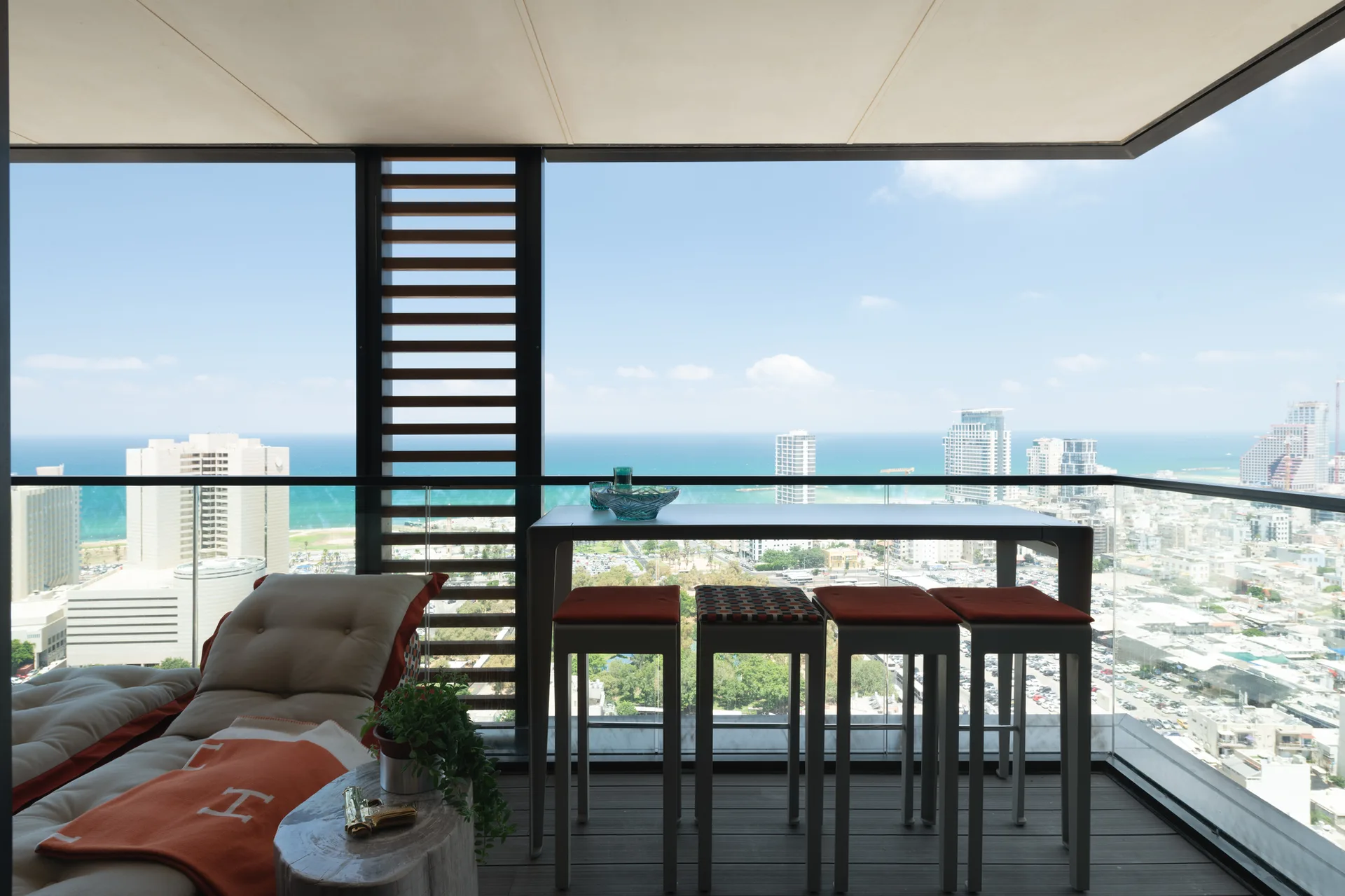 A modern high-rise balcony overlooking the ocean features a slim bar table with stools, a cushioned lounge chair with an orange blanket, and a glass railing.