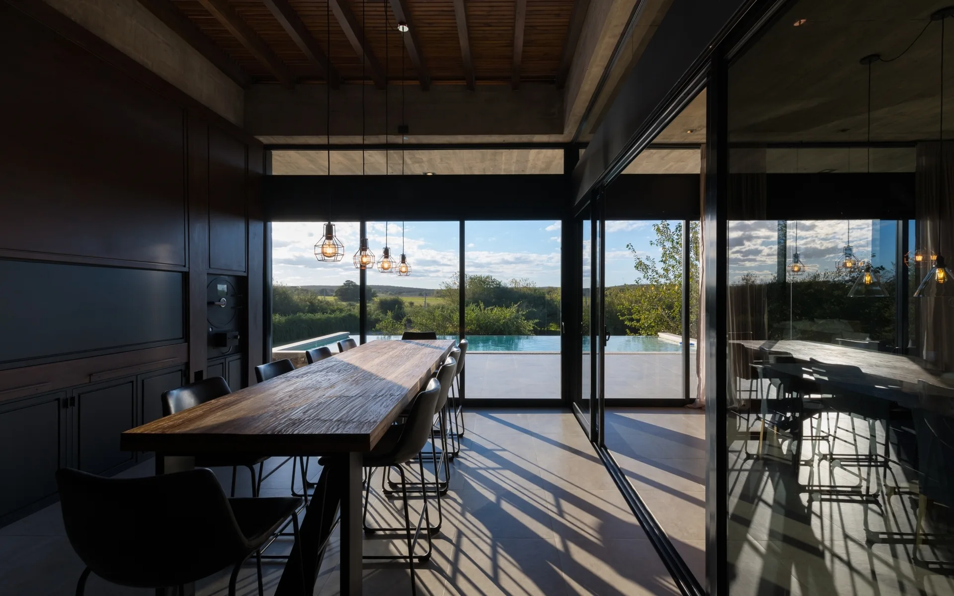 A modern dining room features a long wooden table with dark chairs beneath exposed ceiling beams and glass sliding doors that open to a swimming pool.