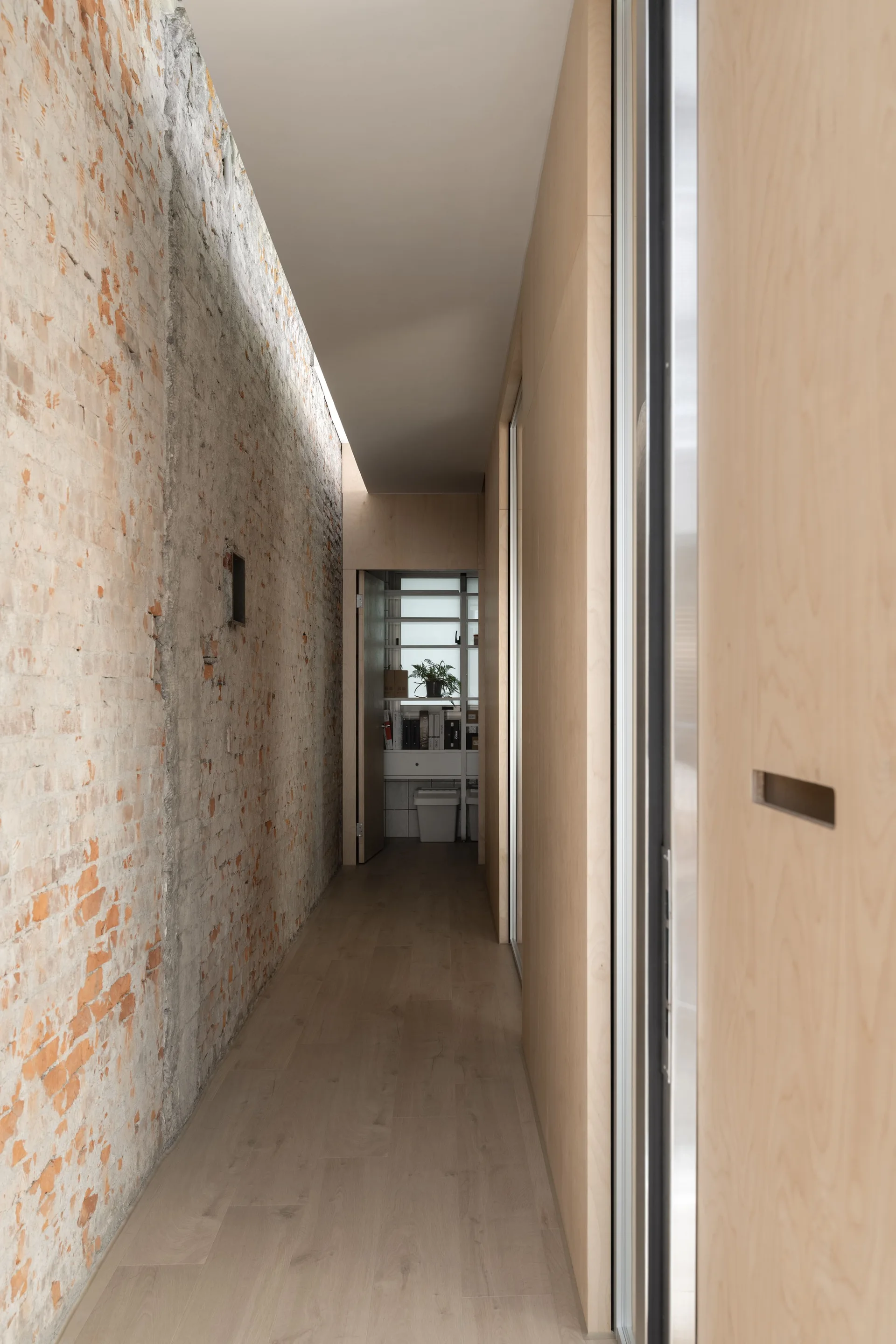 A narrow hallway features a rough exposed brick wall on one side and smooth plywood paneling with light wood flooring on the other.