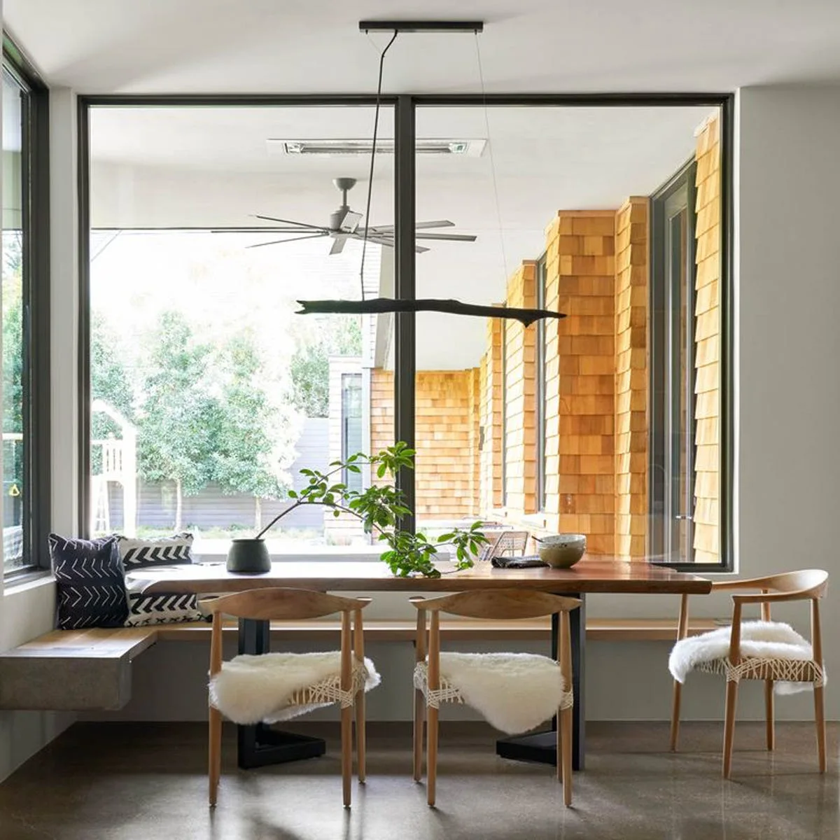 A modern dining nook features a wood table, chairs topped with sheepskins, and a built-in L-shaped bench in front of large windows.