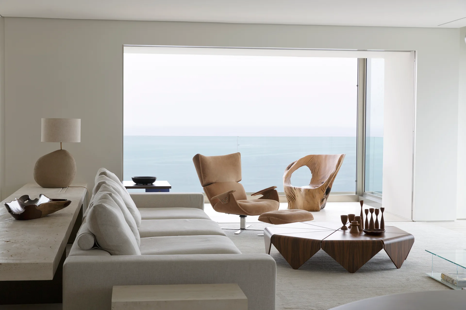 A minimalist living room features a beige sofa, geometric wood coffee table, and sculptural wooden chairs facing a large window with an ocean view.