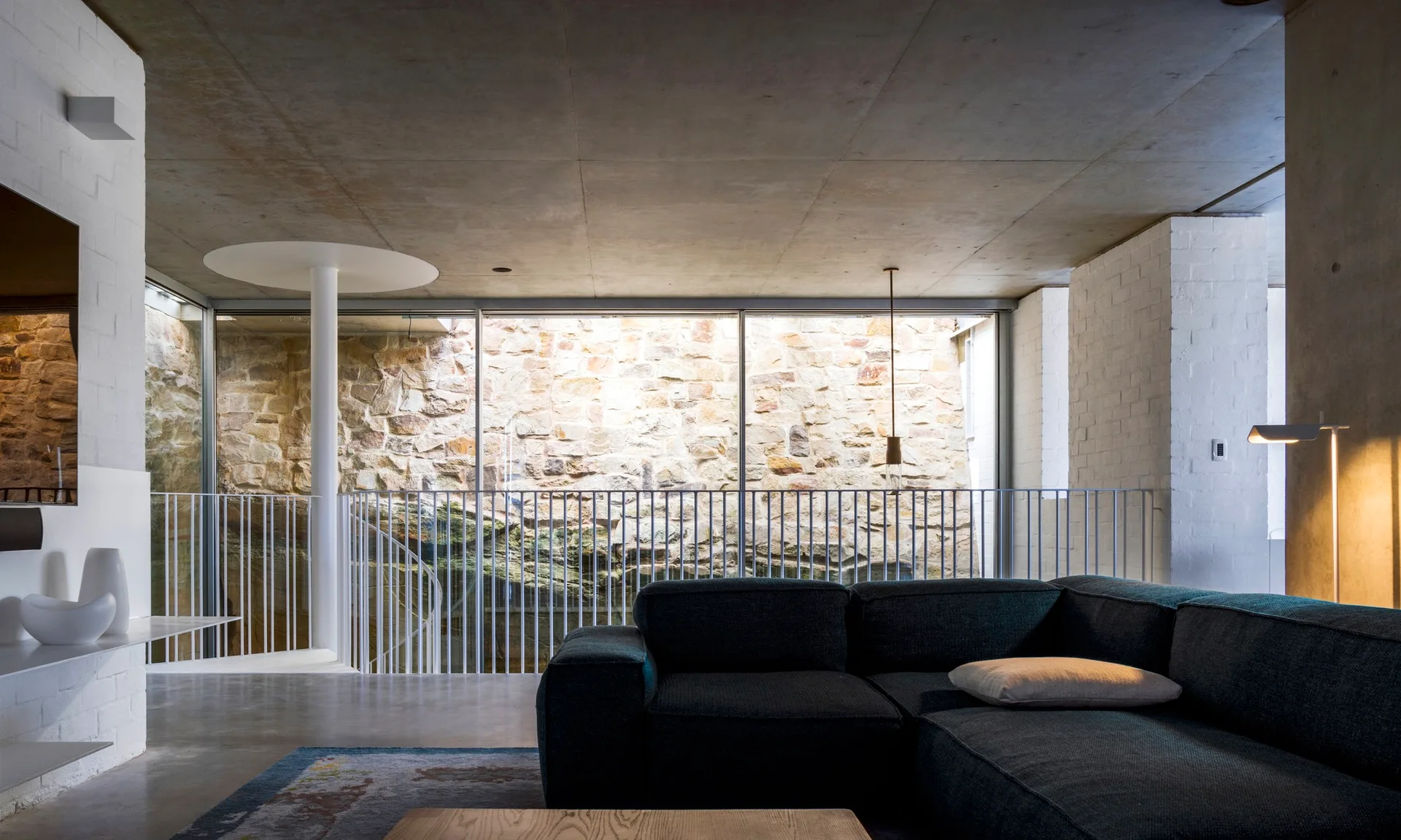 A modern living room features a dark grey sectional sofa, white brick walls, a concrete ceiling, and a glass wall displaying an outdoor stone wall.