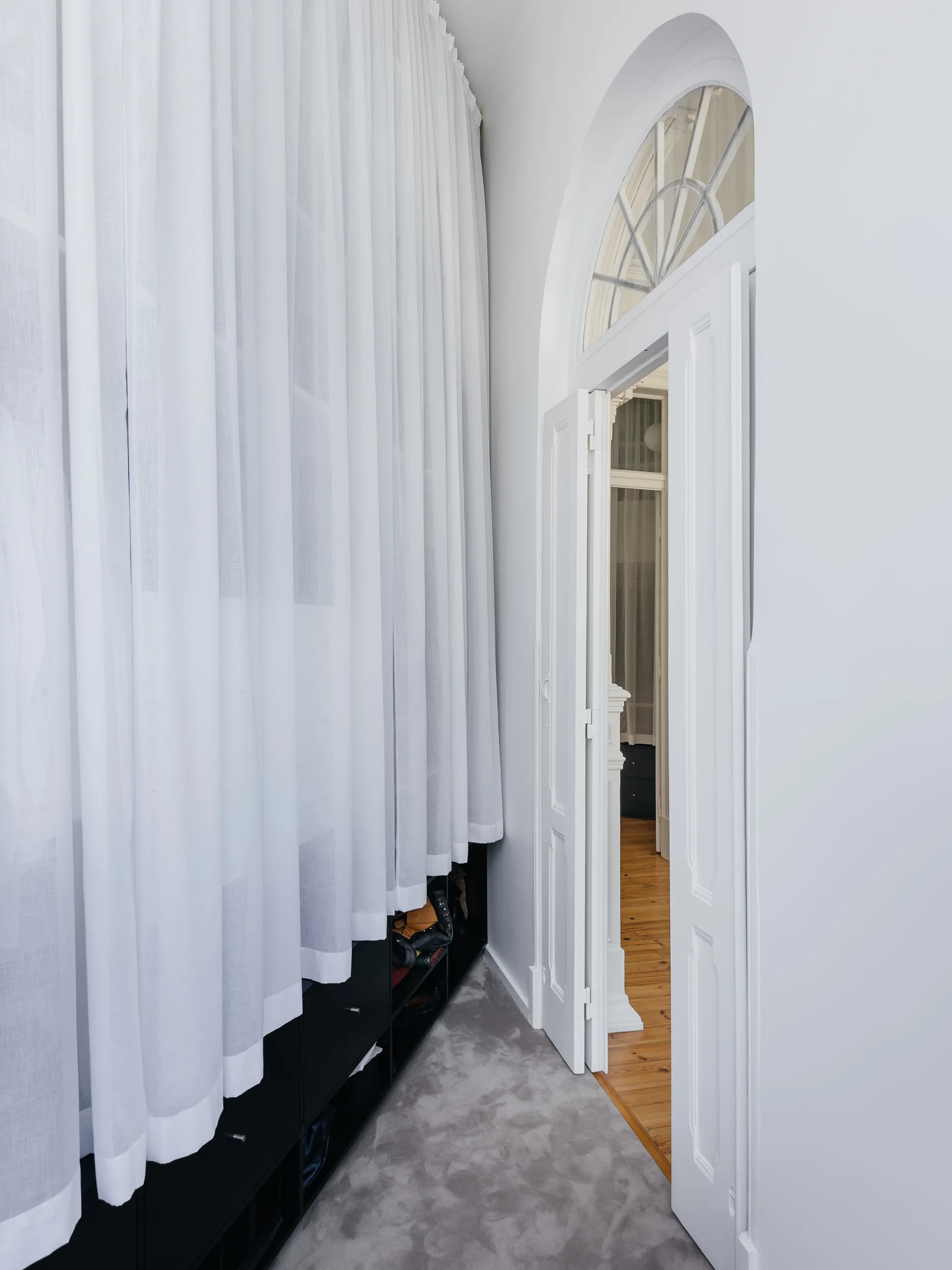 An arched white doorway with a fanlight transom window stands open next to floor-to-ceiling sheer white curtains over a low black storage shelf.