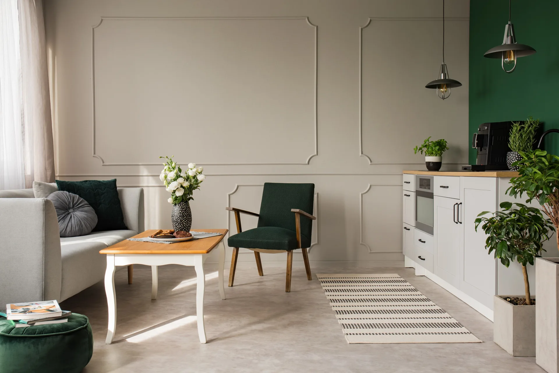 A light grey sofa and dark green armchair sit in a living room with decorative wall molding next to a white kitchen counter under pendant lights.