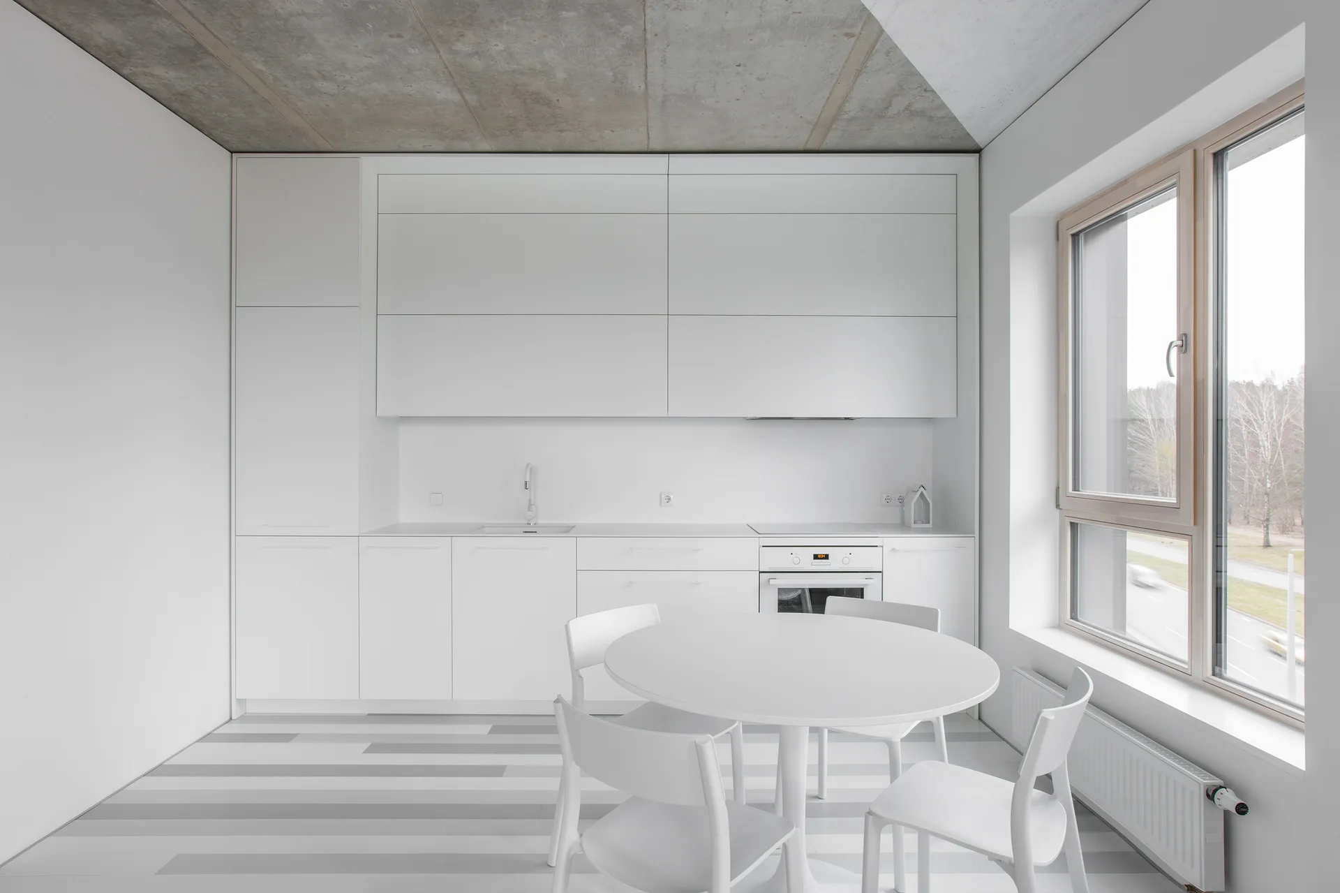 A minimalist white kitchen and dining area features flat-front cabinets, a round table with chairs, a striped floor, and an exposed concrete ceiling.
