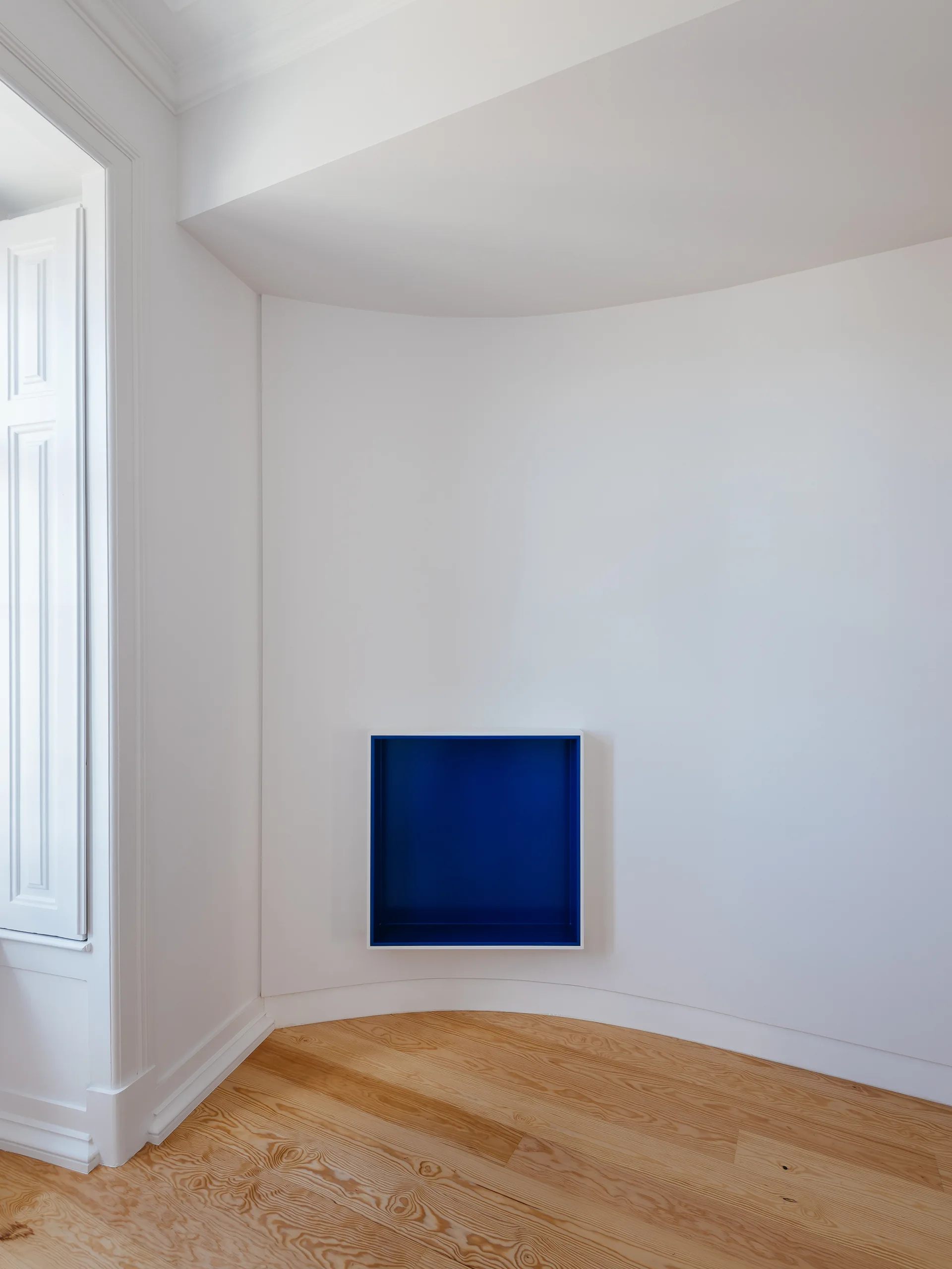 A square cobalt blue recessed wall niche built into a smooth white curved wall above light-toned wood flooring.