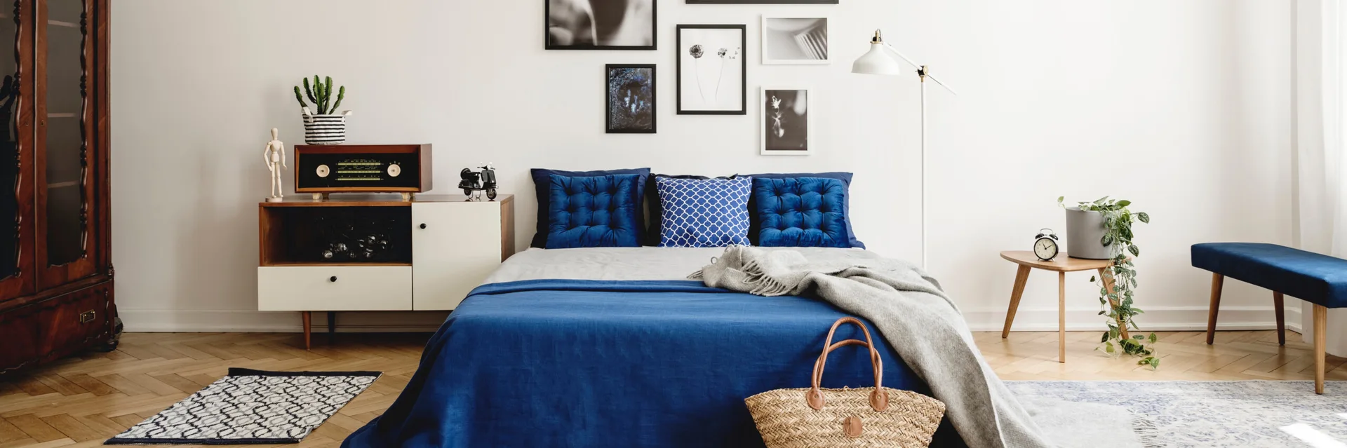 A bright minimalist bedroom features a bed with cobalt blue bedding, a gallery wall of black and white art prints, and a mid-century sideboard.