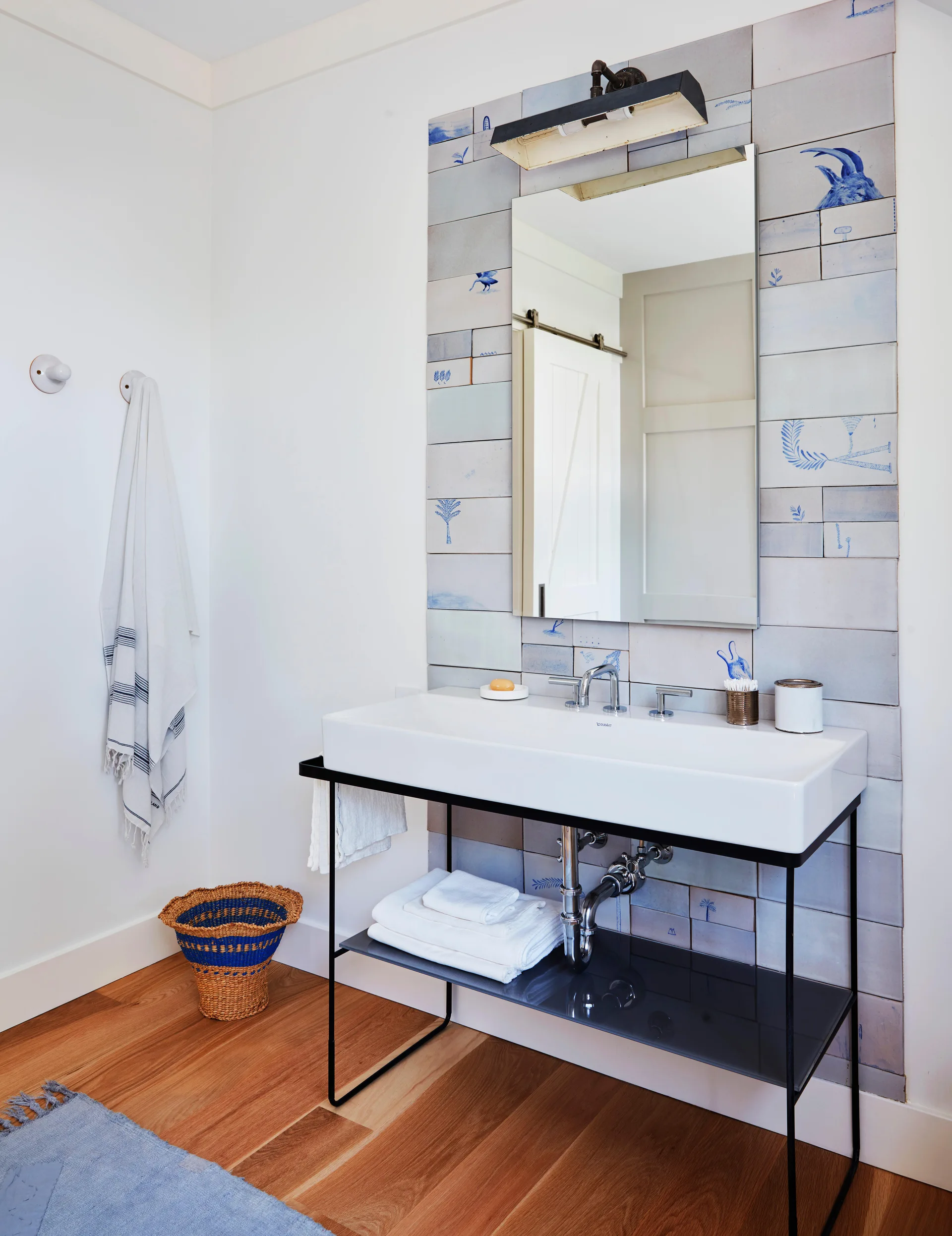 A modern bathroom features a white washbasin with a black metal console frame set against a vertical strip of illustrated blue and white tiles.
