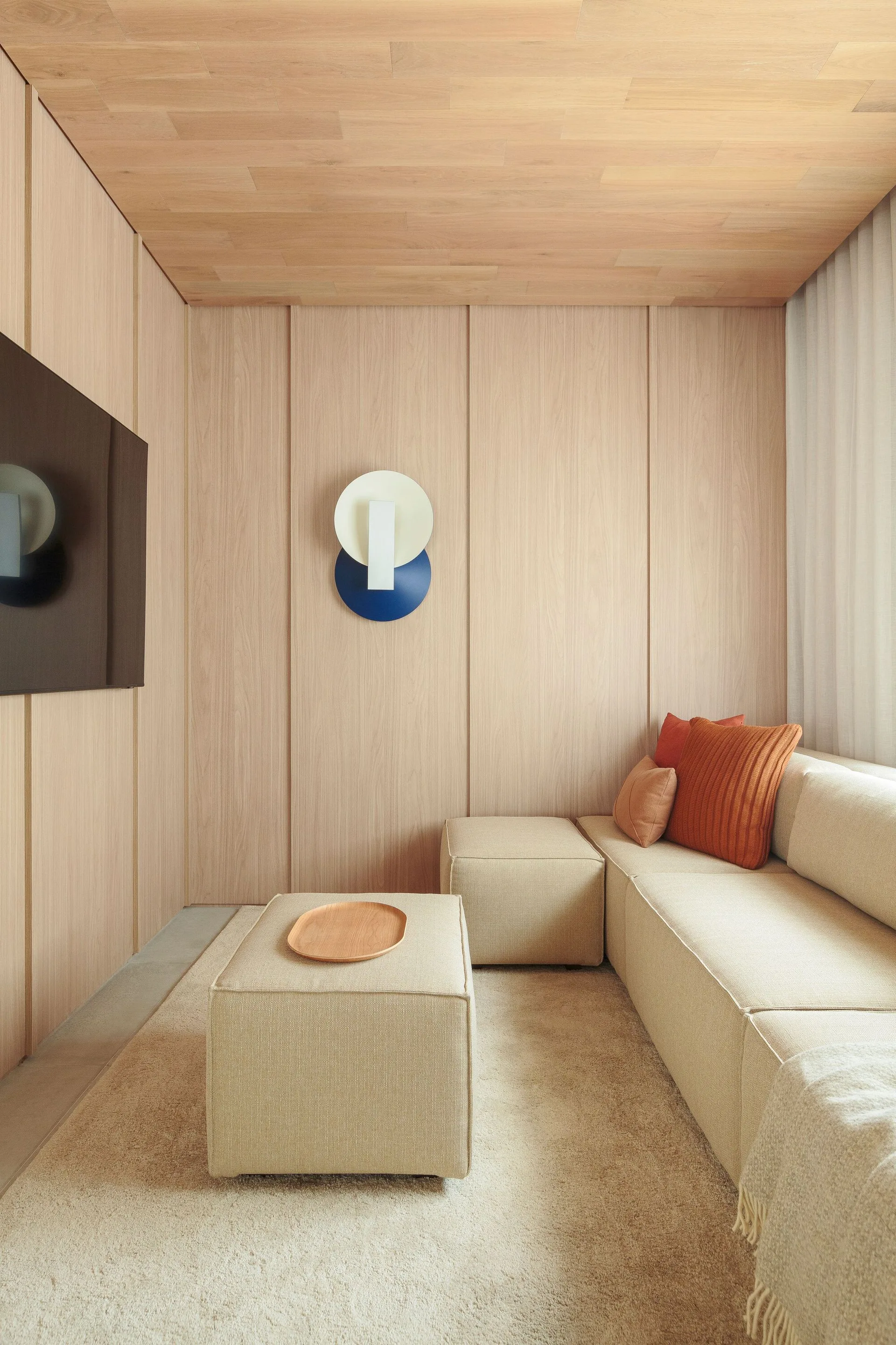 A modern lounge features light wood paneling on the walls and ceiling, a beige modular sectional sofa, and a wall-mounted television.