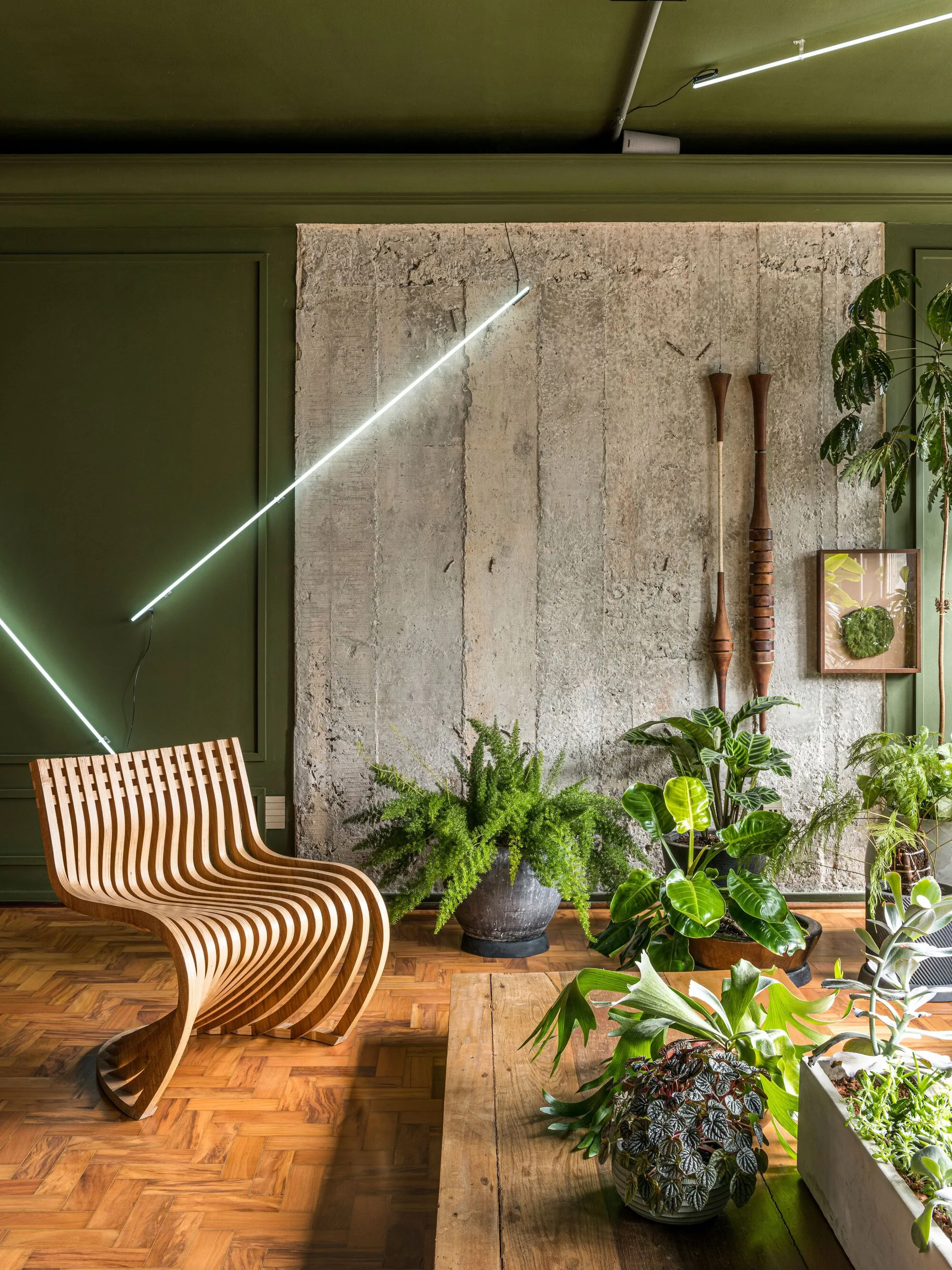 A stylish room featuring a curved slatted wood chair, an exposed concrete accent wall, lush potted houseplants, and a diagonal neon tube light.
