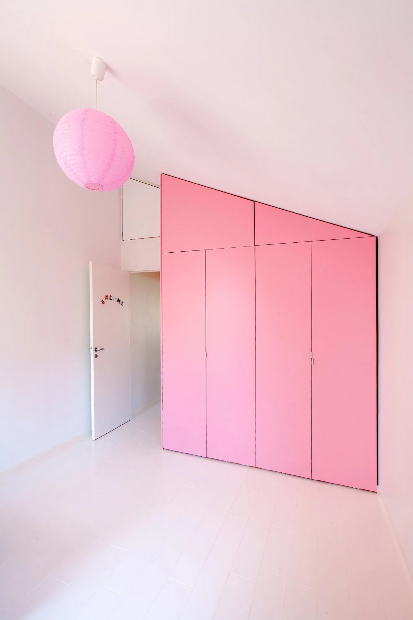 A minimalist white bedroom features a large, custom-built pink wardrobe designed to fit beneath a sloped ceiling next to a matching pink paper lantern.