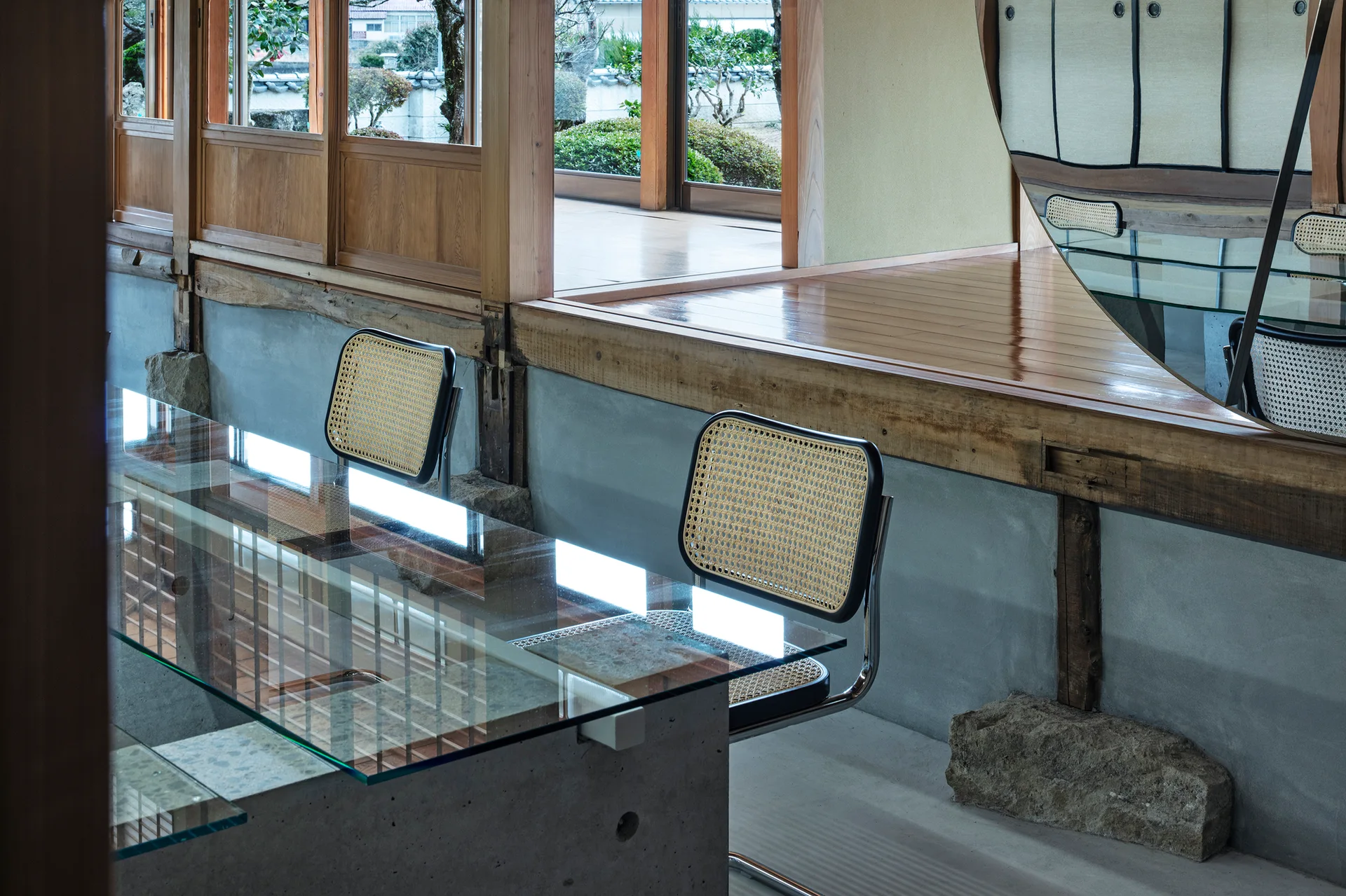 A glass-topped table with concrete bases is paired with cane and chrome chairs next to an elevated wooden platform and sliding window panels.