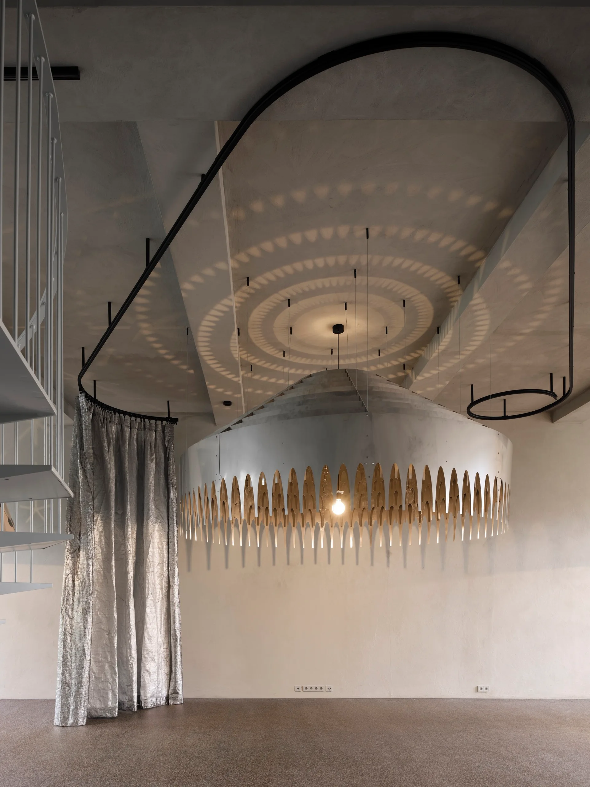 A large scalloped metal pendant light casts concentric shadow circles on a concrete ceiling beside a silver curtain and spiral staircase.