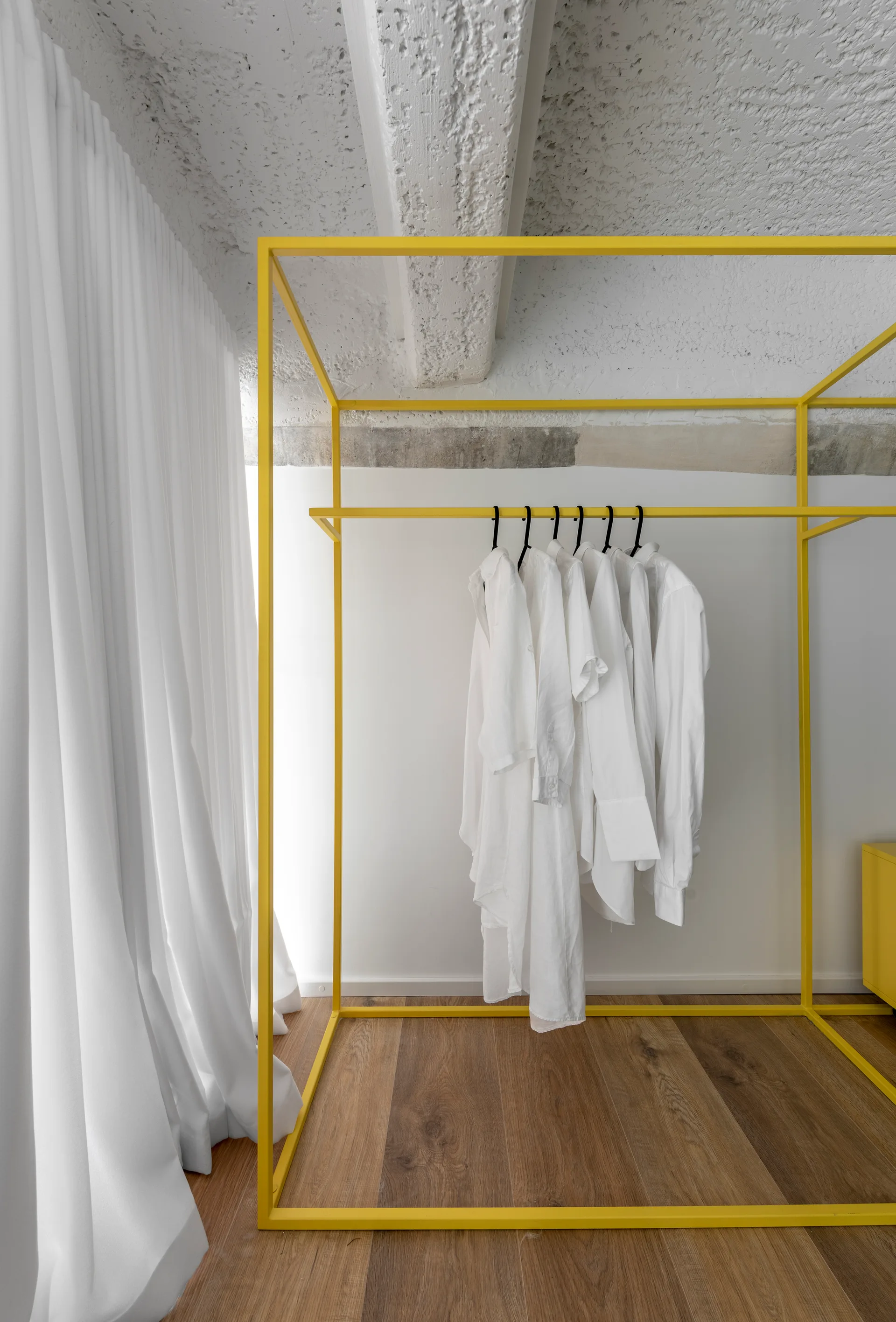 A minimalist bedroom featuring a bright yellow metal clothes rack hanging white shirts, situated on wood flooring next to long white curtains.