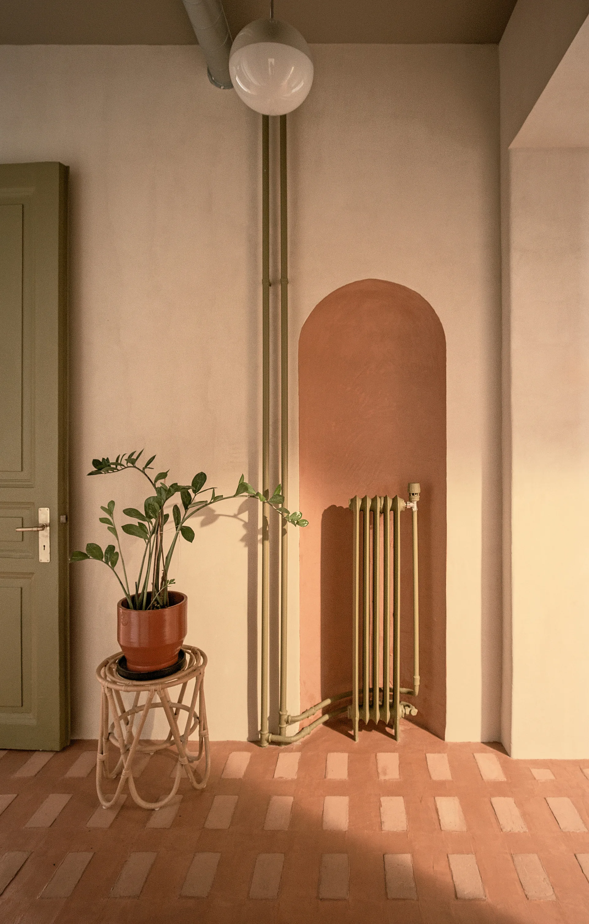 An arched terracotta niche holds a green radiator next to exposed green pipes, a potted plant on a rattan stand, and a green door.