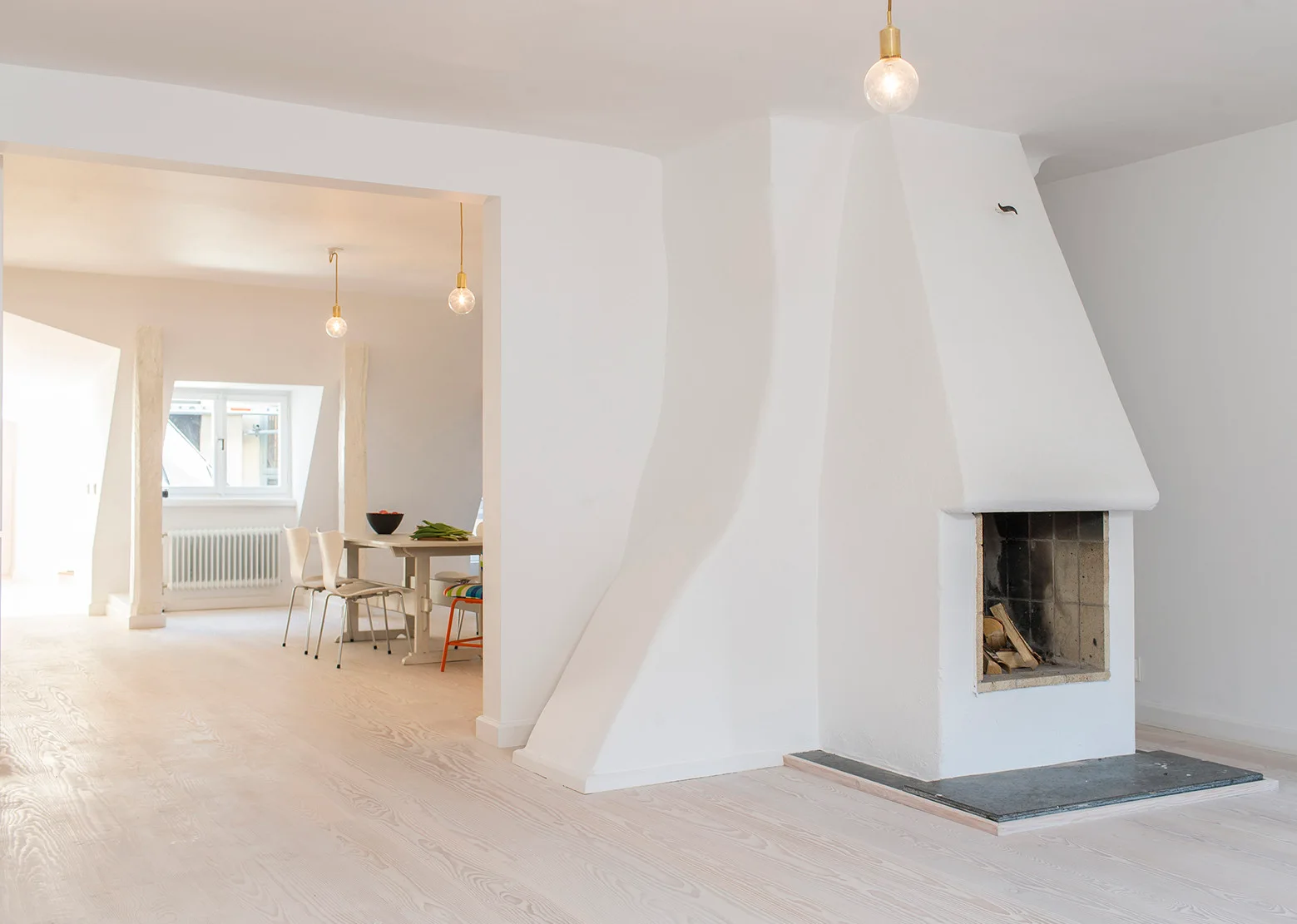 A minimalist living room with a white sculptural fireplace, white-washed wood floors, and an opening that leads to a dining area.