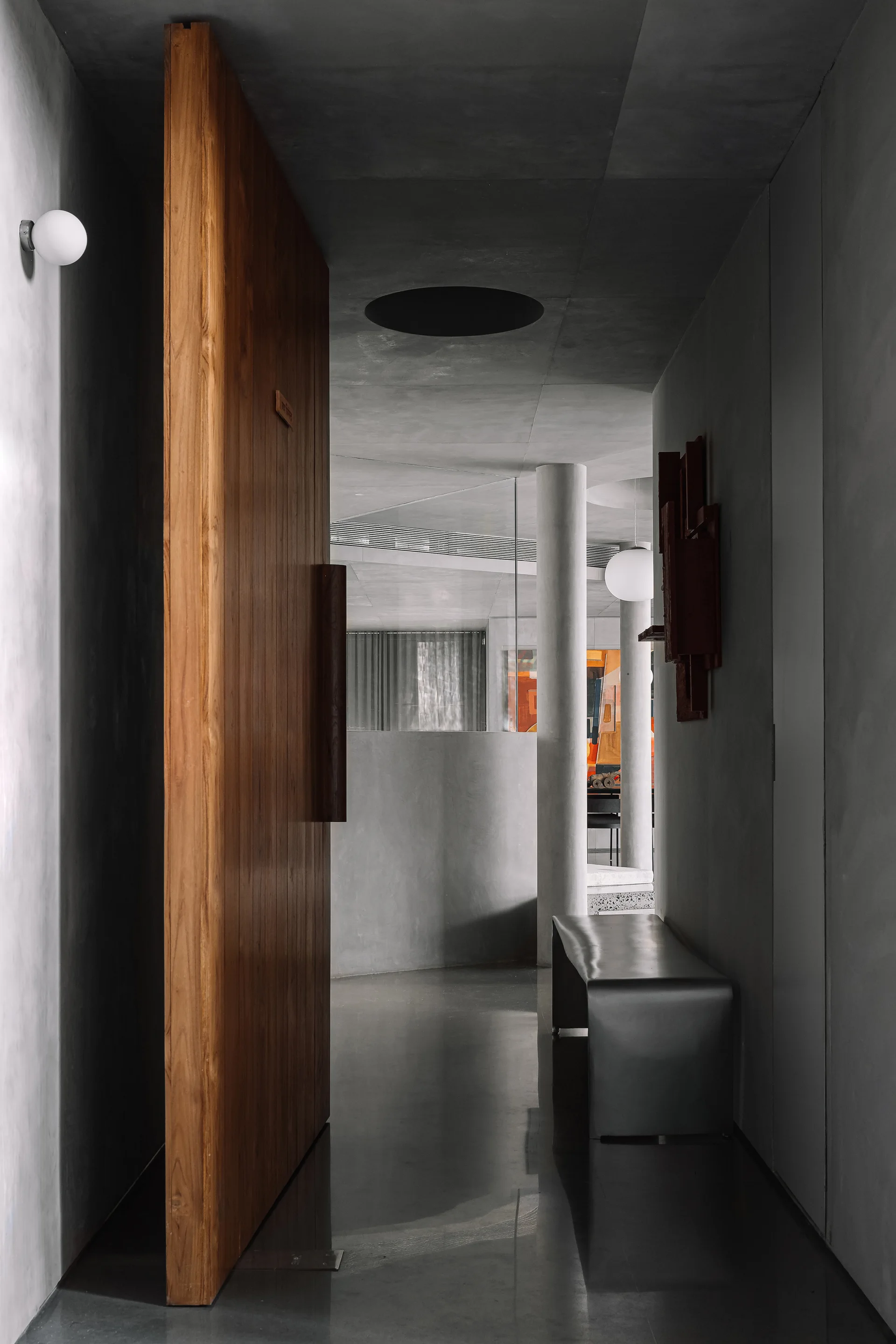 A minimalist concrete corridor features a massive wooden pivot door, polished grey flooring, a simple metal bench, and a round wall sconce.