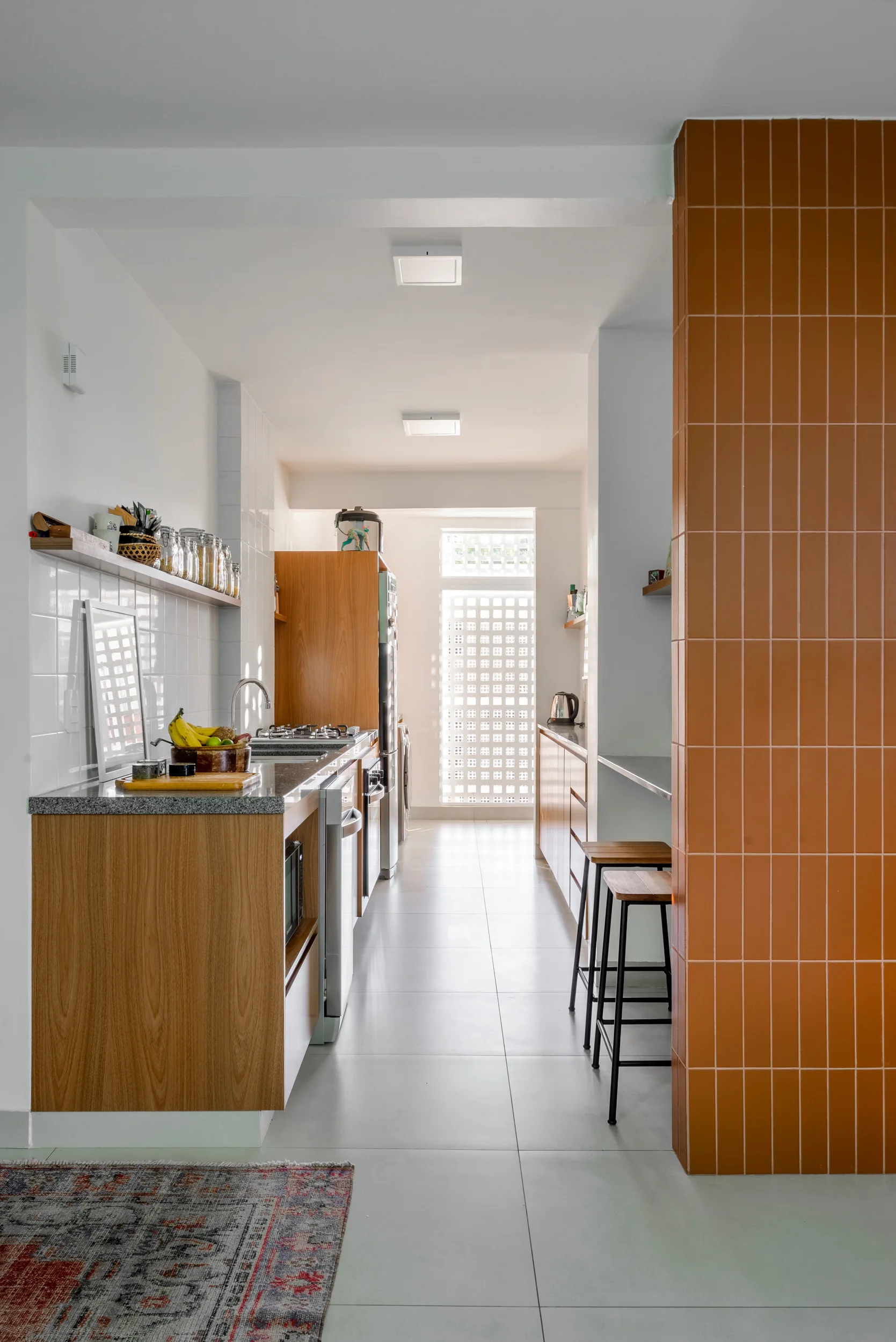 A modern galley kitchen featuring light wood cabinets, gray countertops, a vertical orange tiled column, and wooden bar stools.