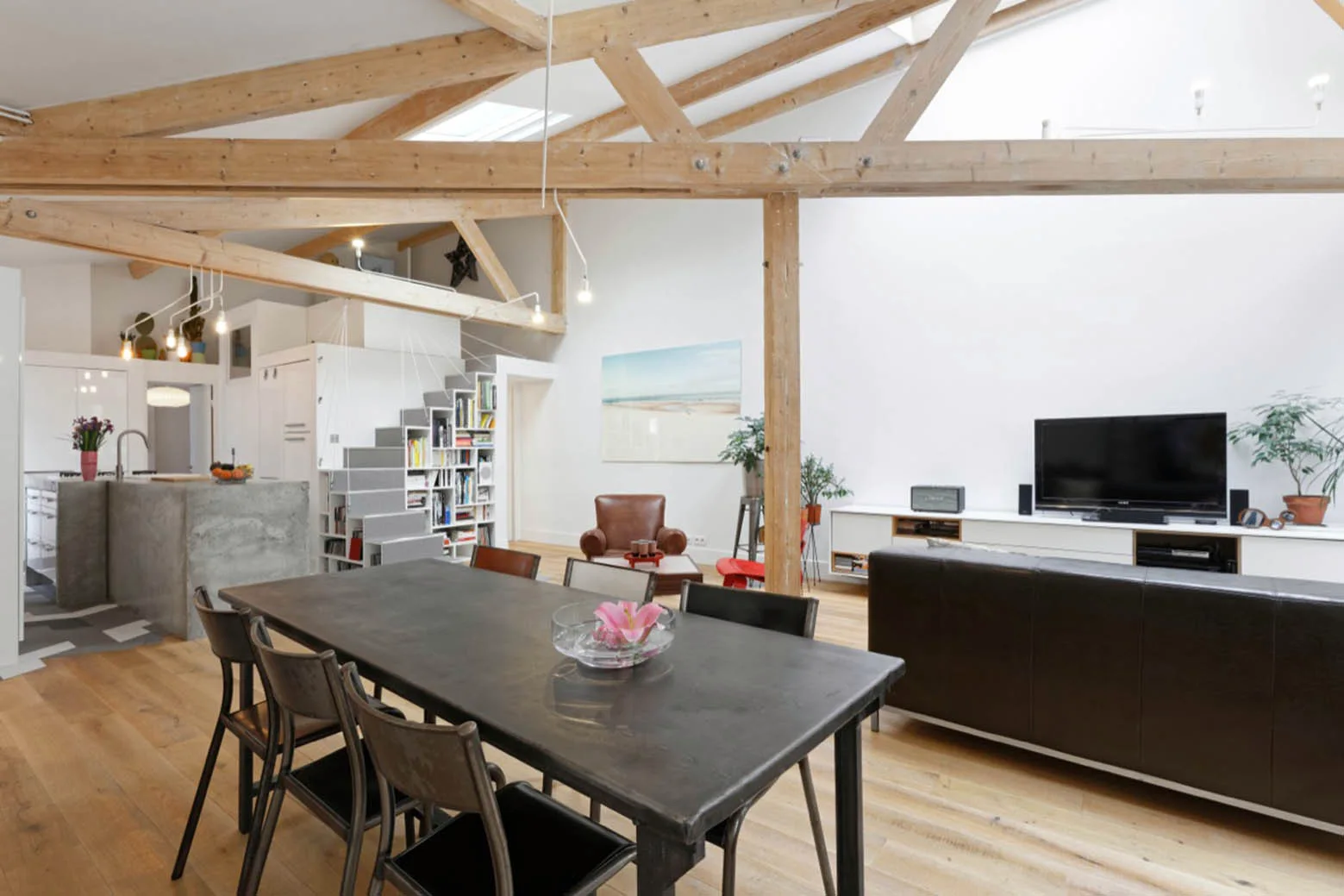 An open-plan loft living room features exposed timber ceiling beams, a dark wood dining table, a brown leather sofa, and a staircase with stepped shelving.