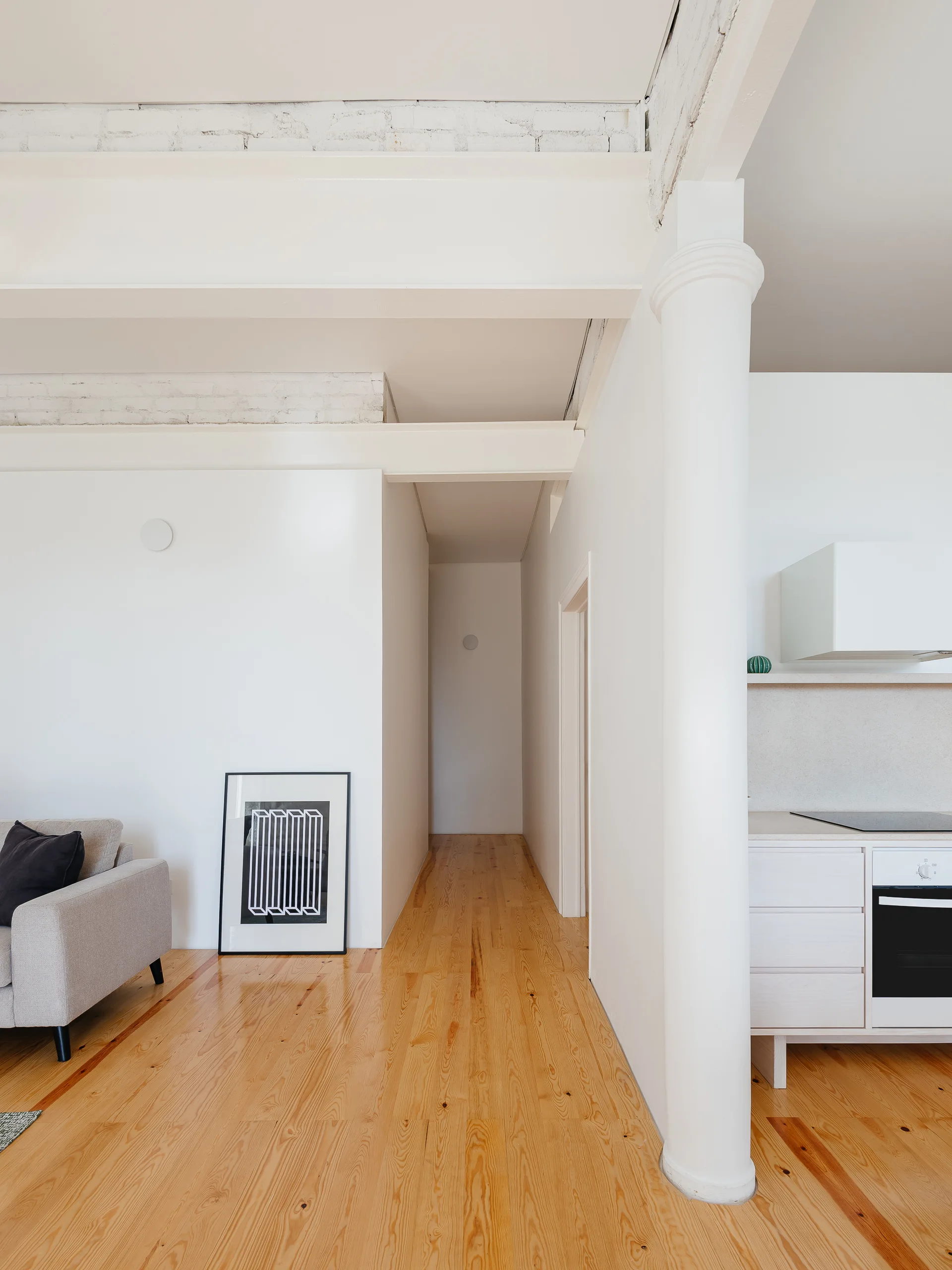 A minimalist apartment interior features a white structural column, natural pine wood floors, a hallway, and a clean white kitchen area.