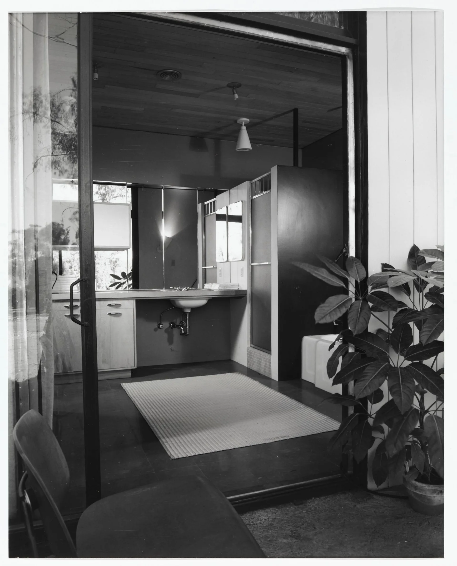 A black and white view through a sliding glass door into a bathroom with a wood plank ceiling, vanity sink, and large mirror.