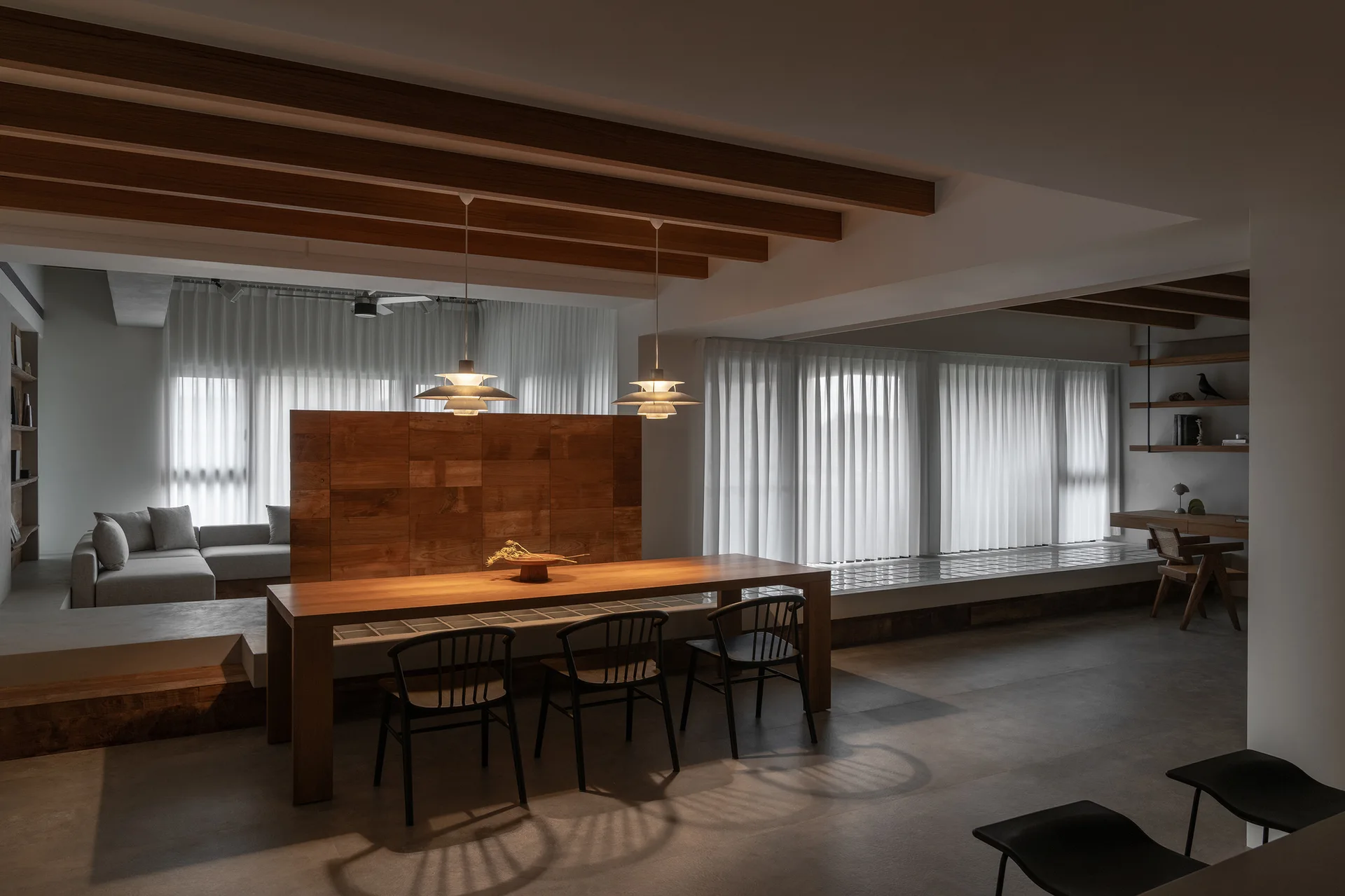 A minimalist dining room with a long wooden table, black chairs, a wood room divider, and a sunken living room in the background.