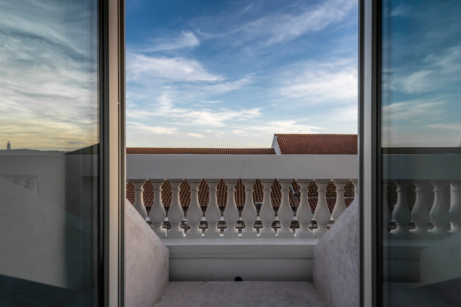 A view through an open glass doorway looking out at a stone balcony with a white balustrade and red tiled rooftops under a blue sky.