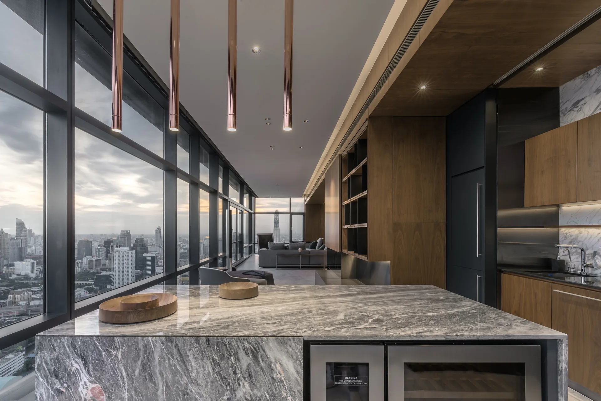 A modern kitchen featuring a gray marble waterfall island, metallic copper pendant lights, and large windows overlooking a city at dusk.