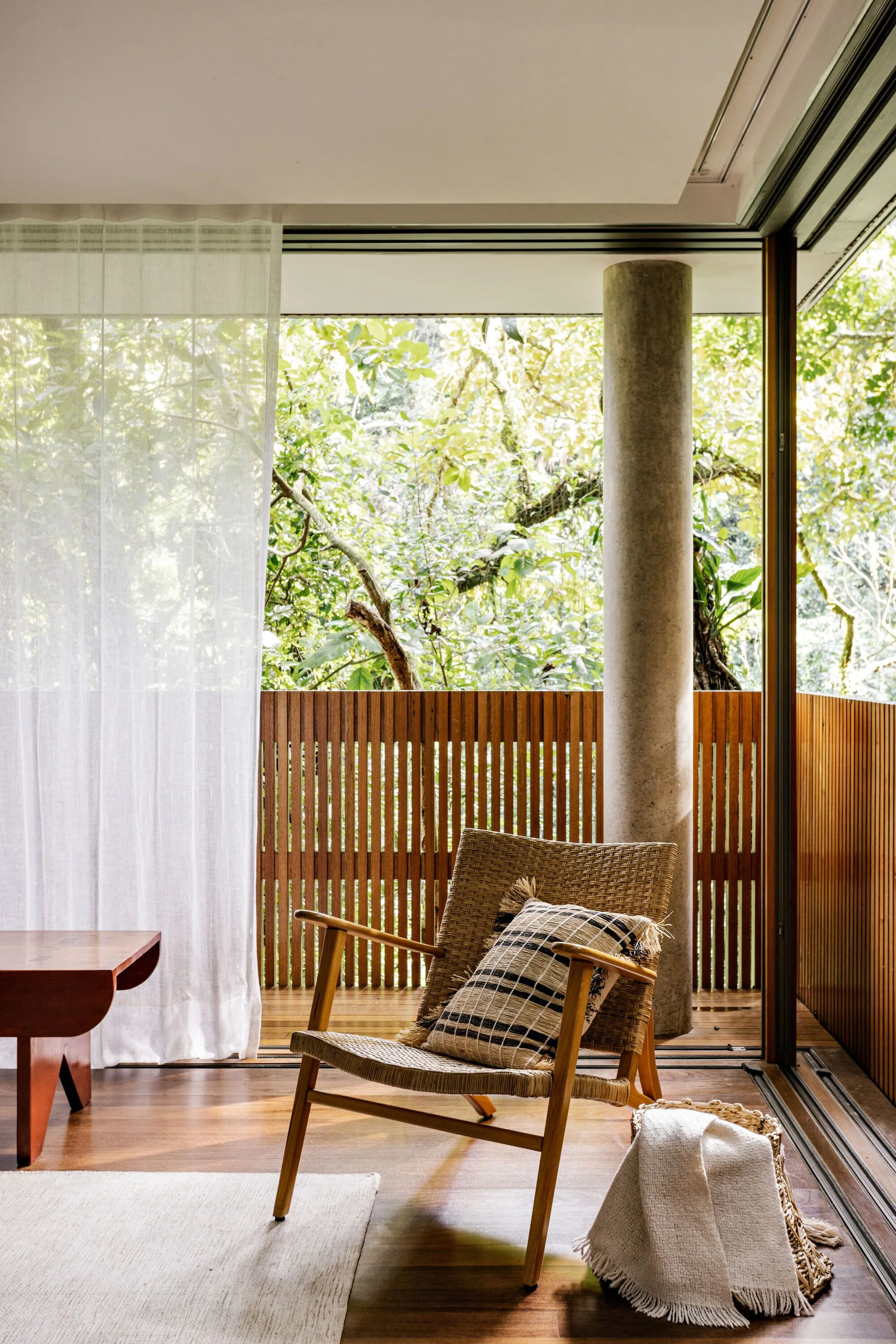 A semi-covered wood patio with a woven lounge chair, a sheer white curtain, a concrete column, and a slatted railing overlooking lush green trees.