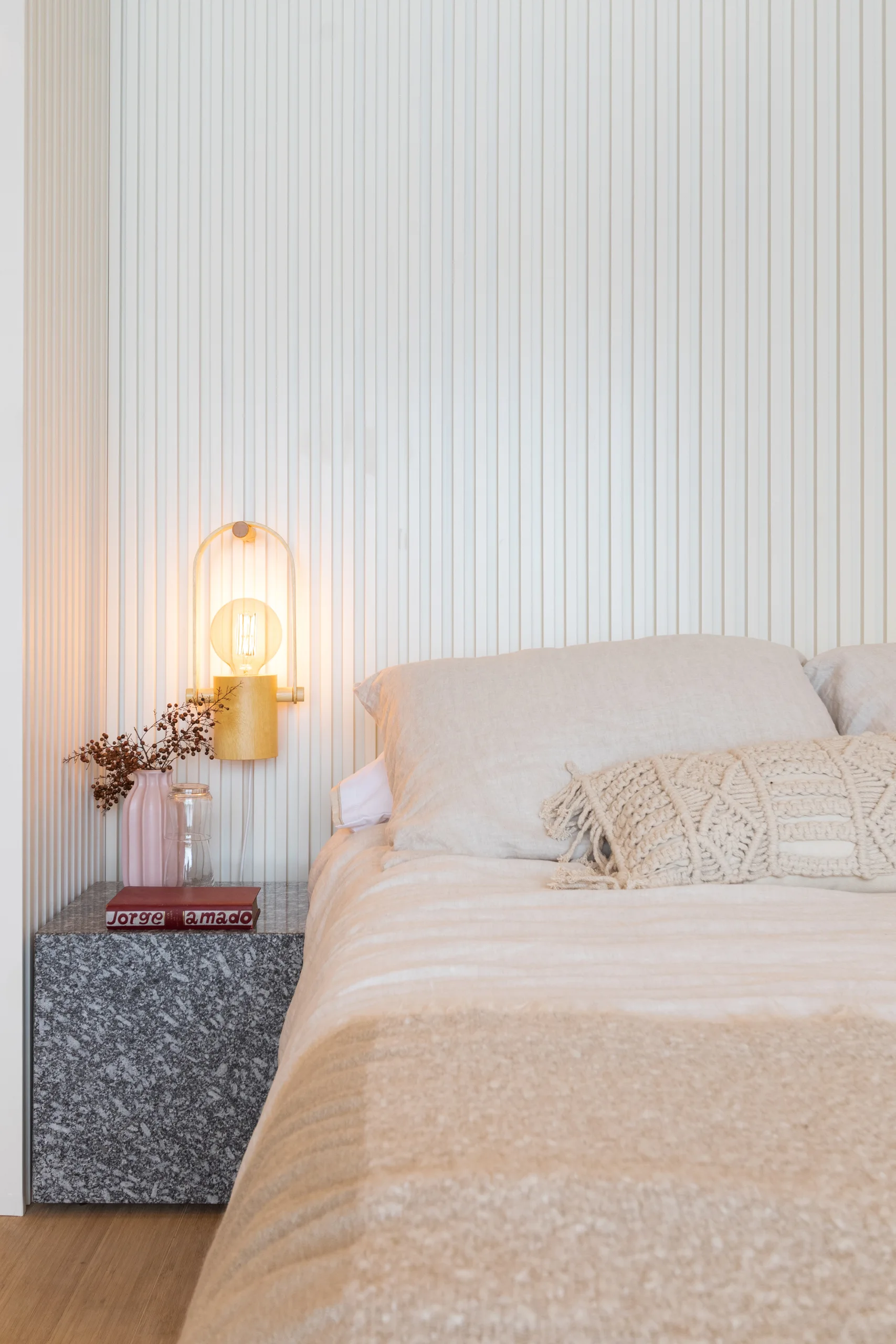 A bedroom features beige linen bedding on the bed next to a speckled stone nightstand and wall lamp against a vertical white slatted wall.