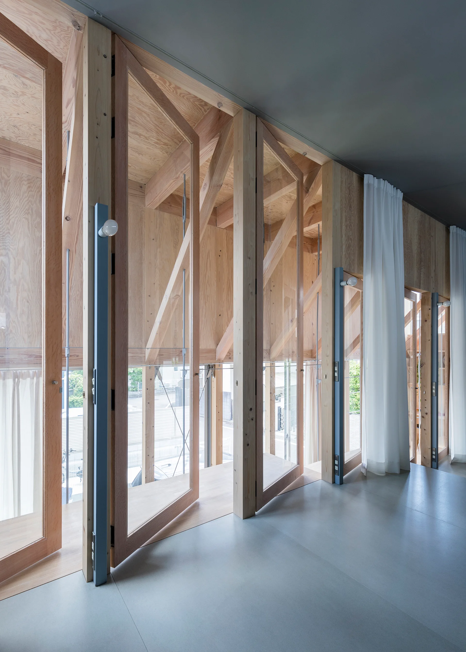 A modern hallway features a wall of operable, hinged glass doors with light wood frames, set against plywood walls and a smooth grey floor.