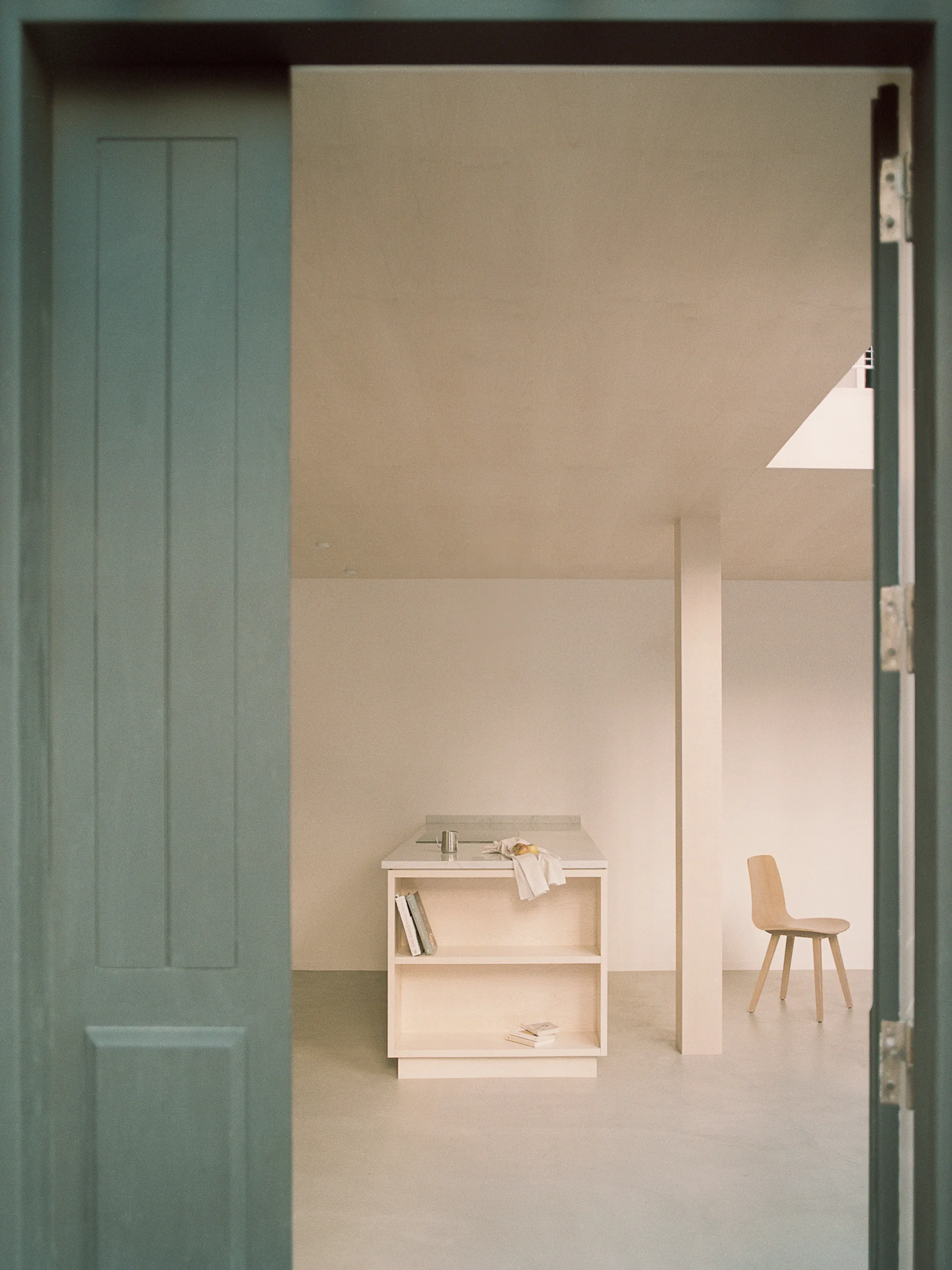 A view through a dark green door frame reveals a minimalist beige kitchen with a wooden island, open shelving, and a single wooden chair.
