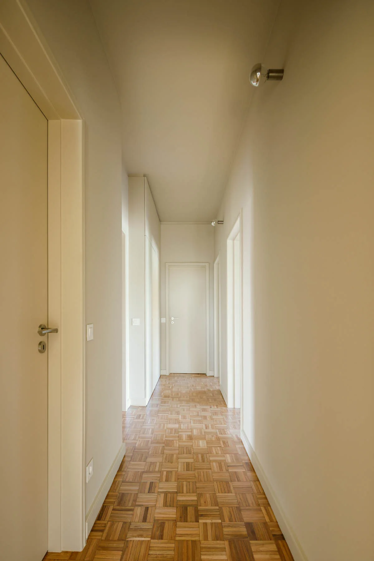 A long minimalist hallway with cream walls, matching painted doors, and a decorative square block wood parquet floor.