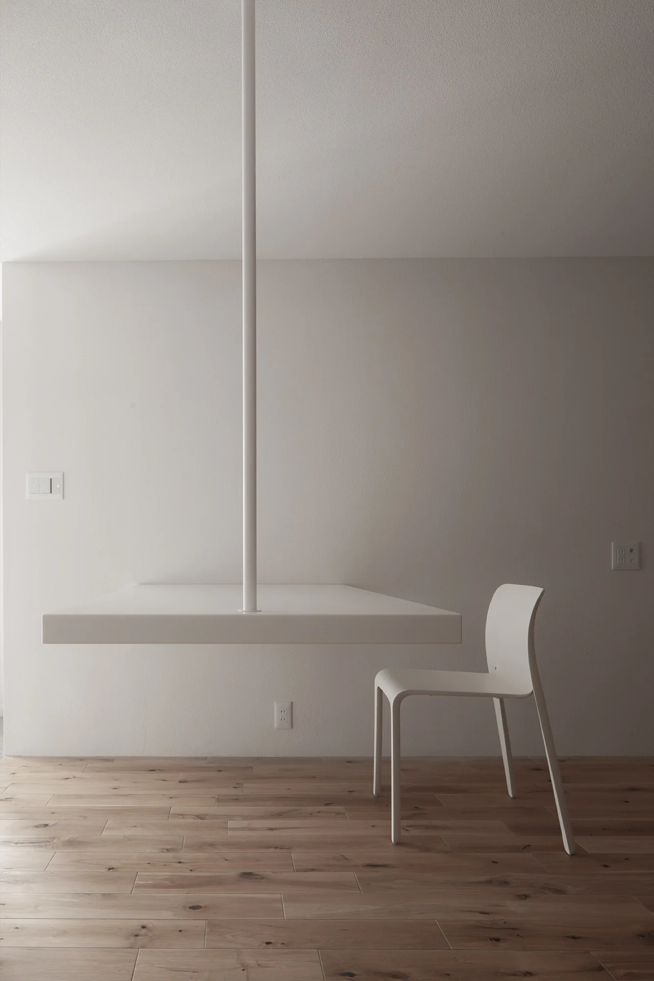 A minimalist white floating desk is suspended from the ceiling by a vertical rod next to a simple white chair on light-toned wood floors.