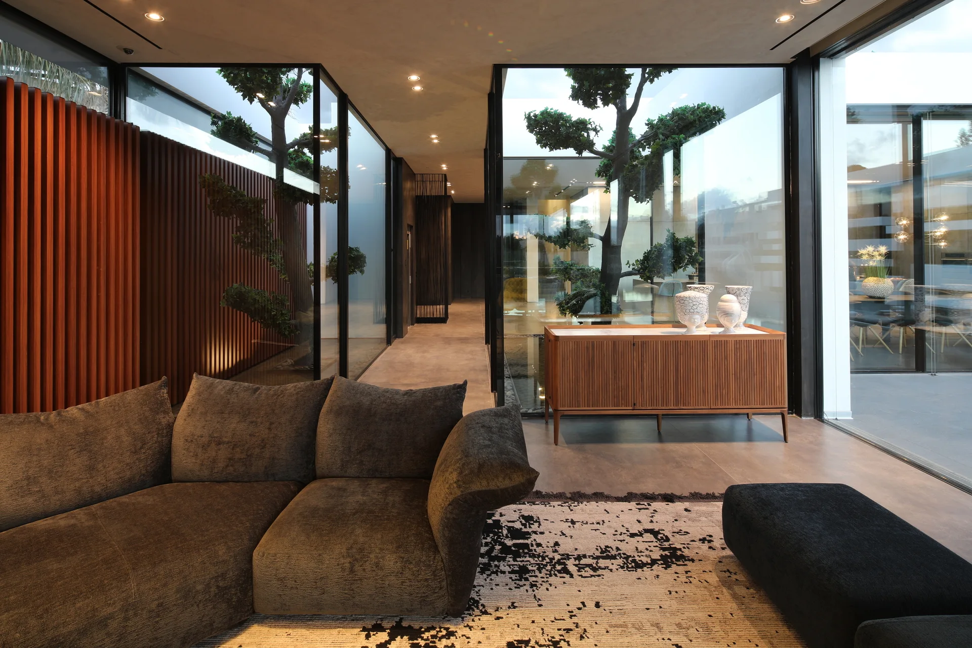 A modern living room features a grey sofa, patterned rug, and a slatted wood console next to glass walls showcasing an outdoor courtyard with a tree.