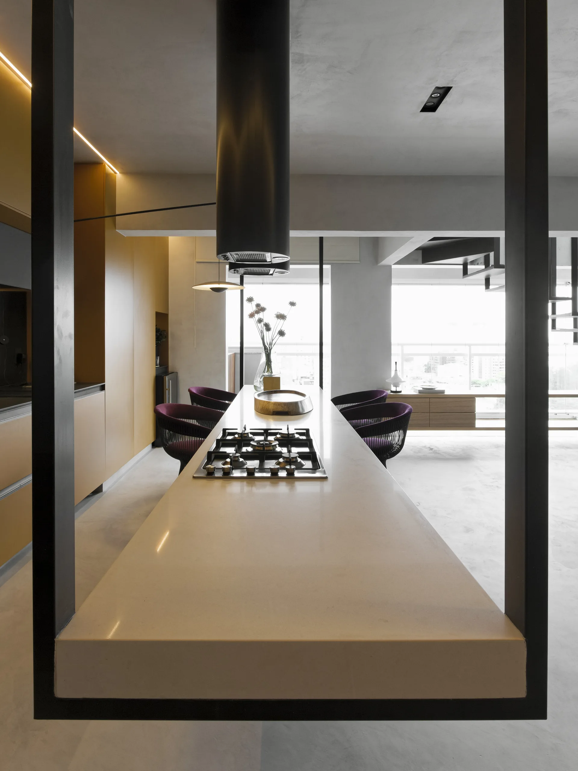 A long minimalist kitchen island features an integrated cooktop, a black cylindrical range hood, deep purple chairs, and ochre cabinets in the background.
